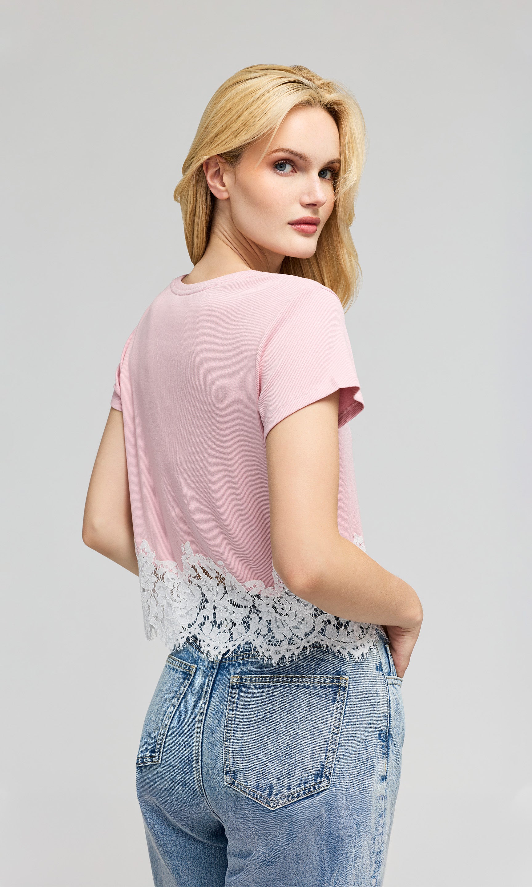 Back view of woman wearing a pink top with lace details and blue jeans on a gray background