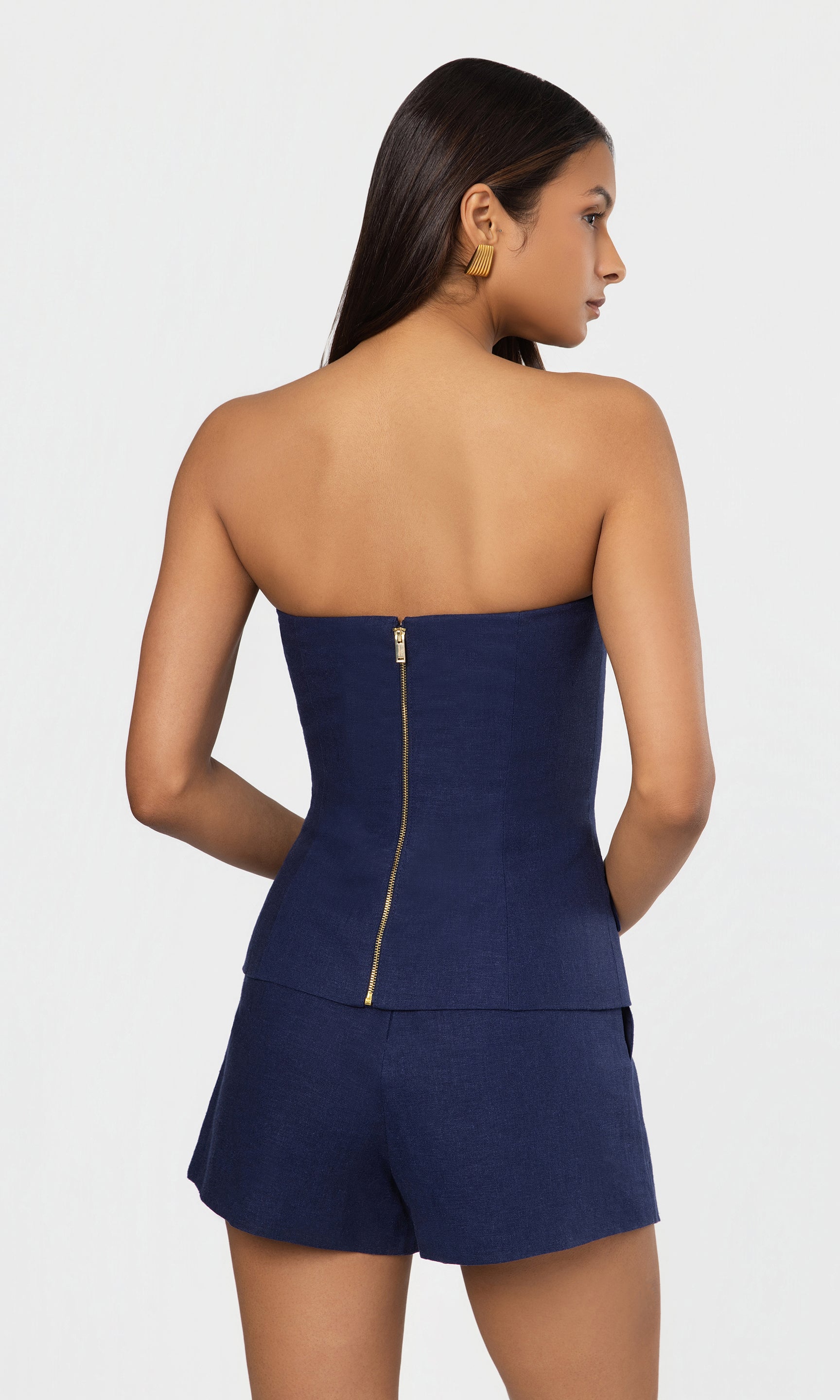 Back view of woman wearing a strapless navy romper with a gold zipper on a white background