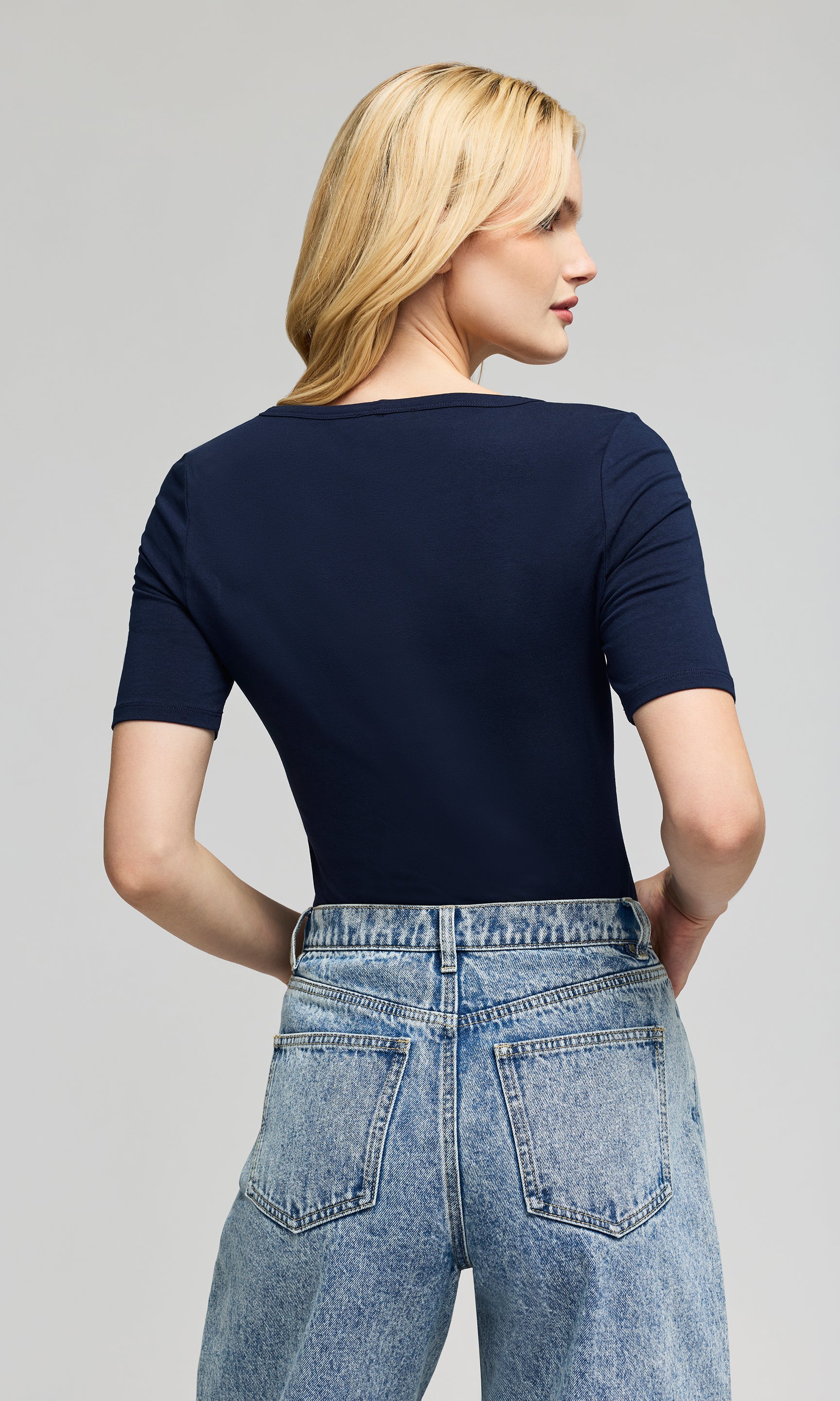 Back view of woman wearing a navy blue top and light blue jeans on a gray background