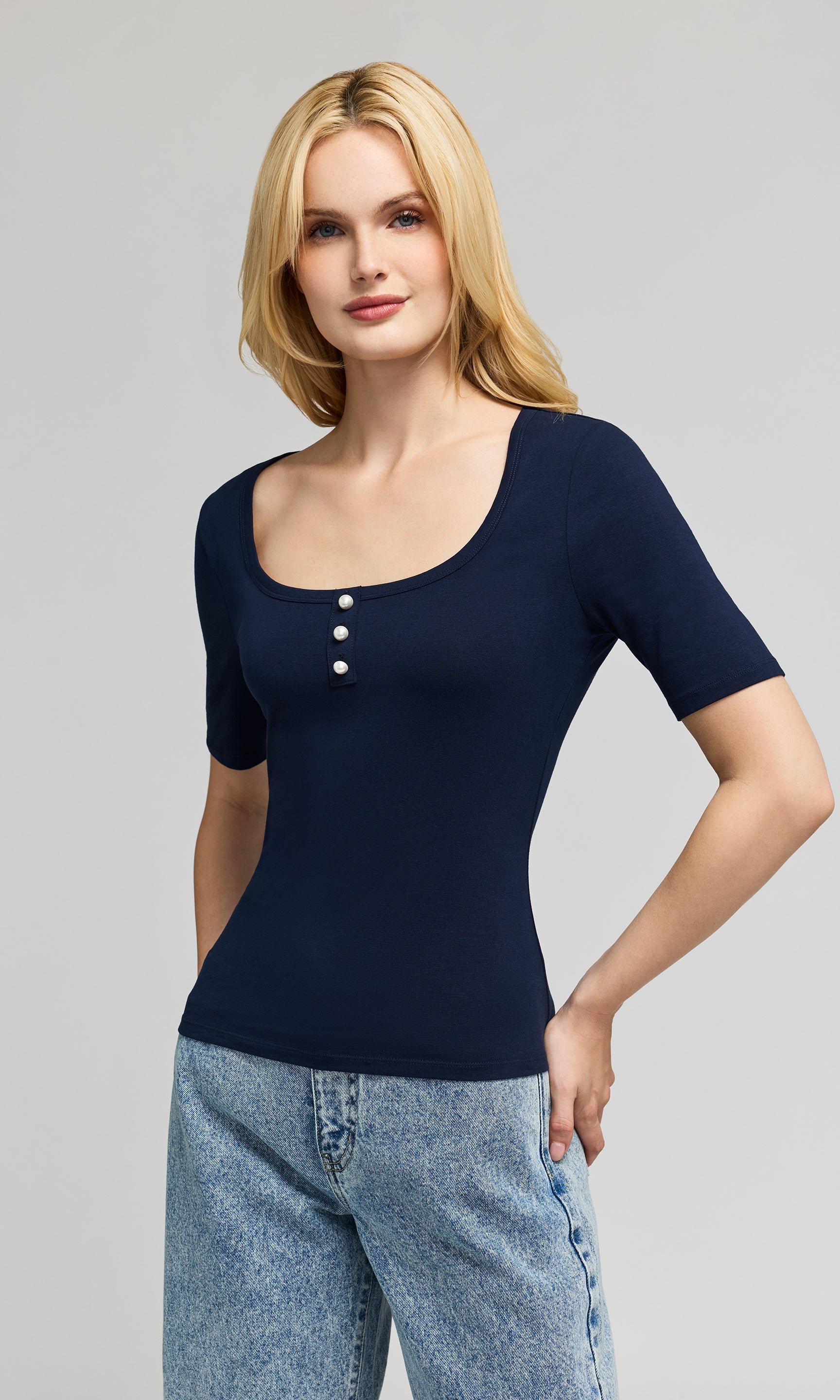 Side view of woman wearing a navy blue short-sleeve top with pearl buttons and light blue jeans on a gray background