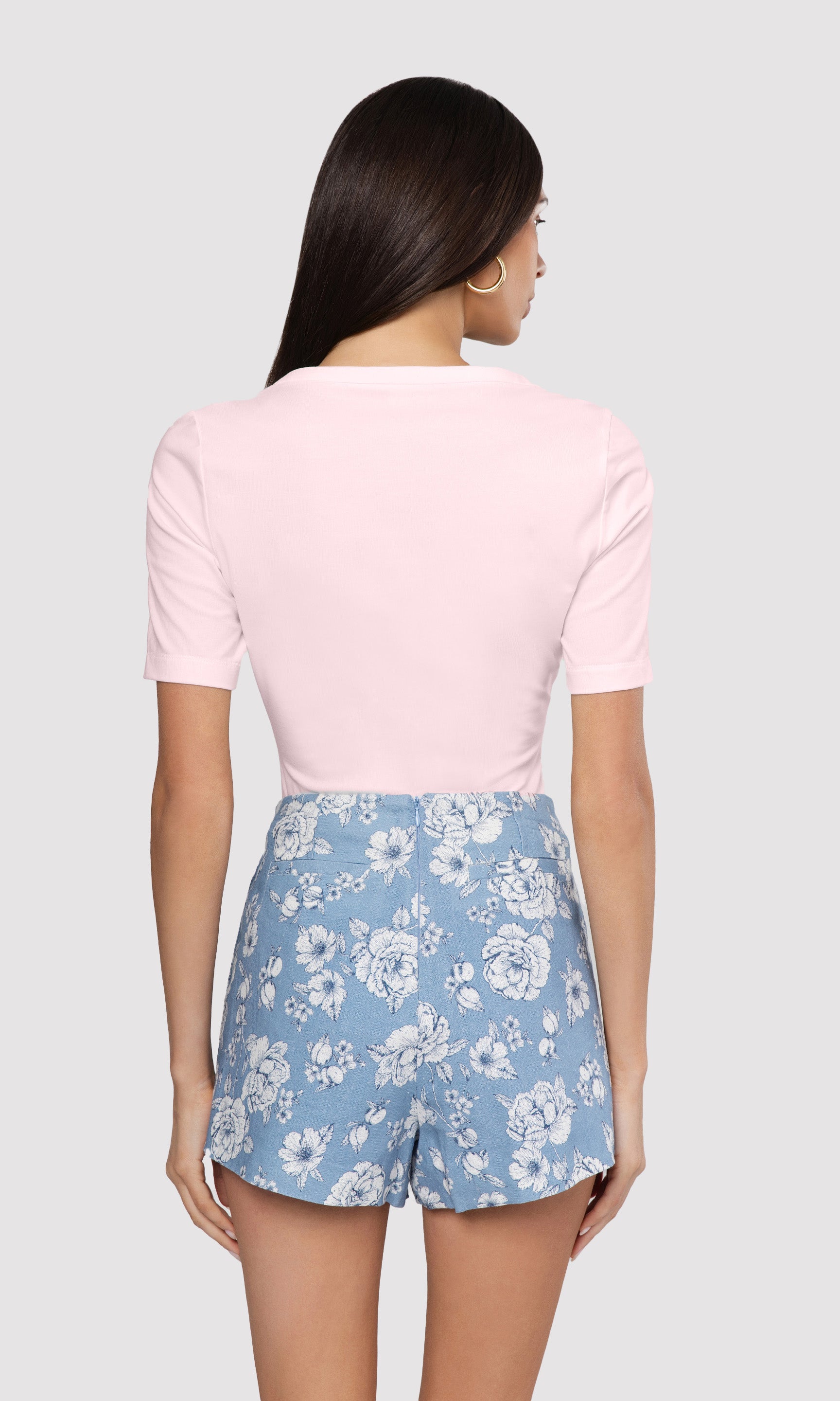 Back view of woman wearing a pink top and blue floral shorts on a white background
