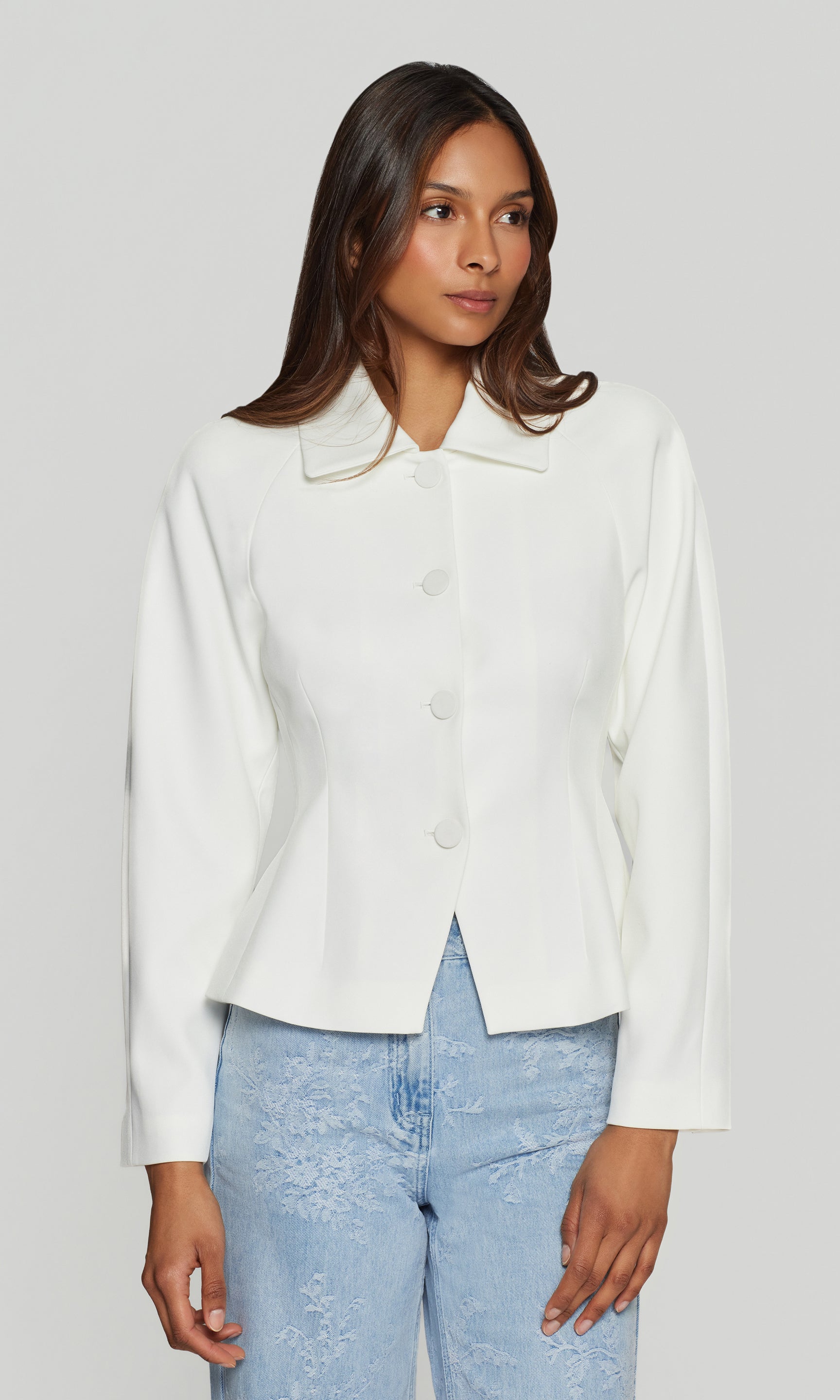 Woman wearing a white jacket and light blue jeans on a plain background