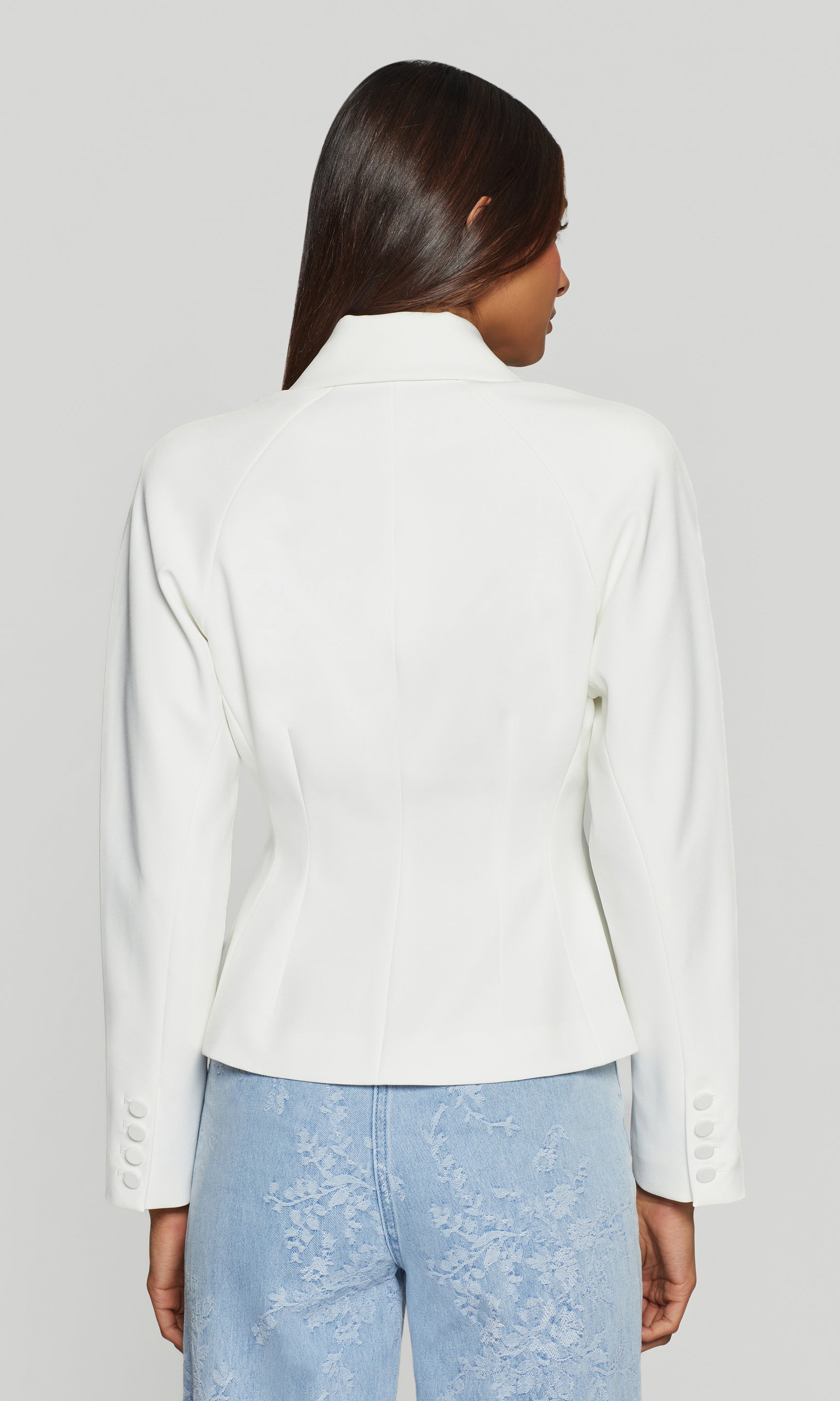 Back view of person wearing a white blazer and light blue pants on a plain background