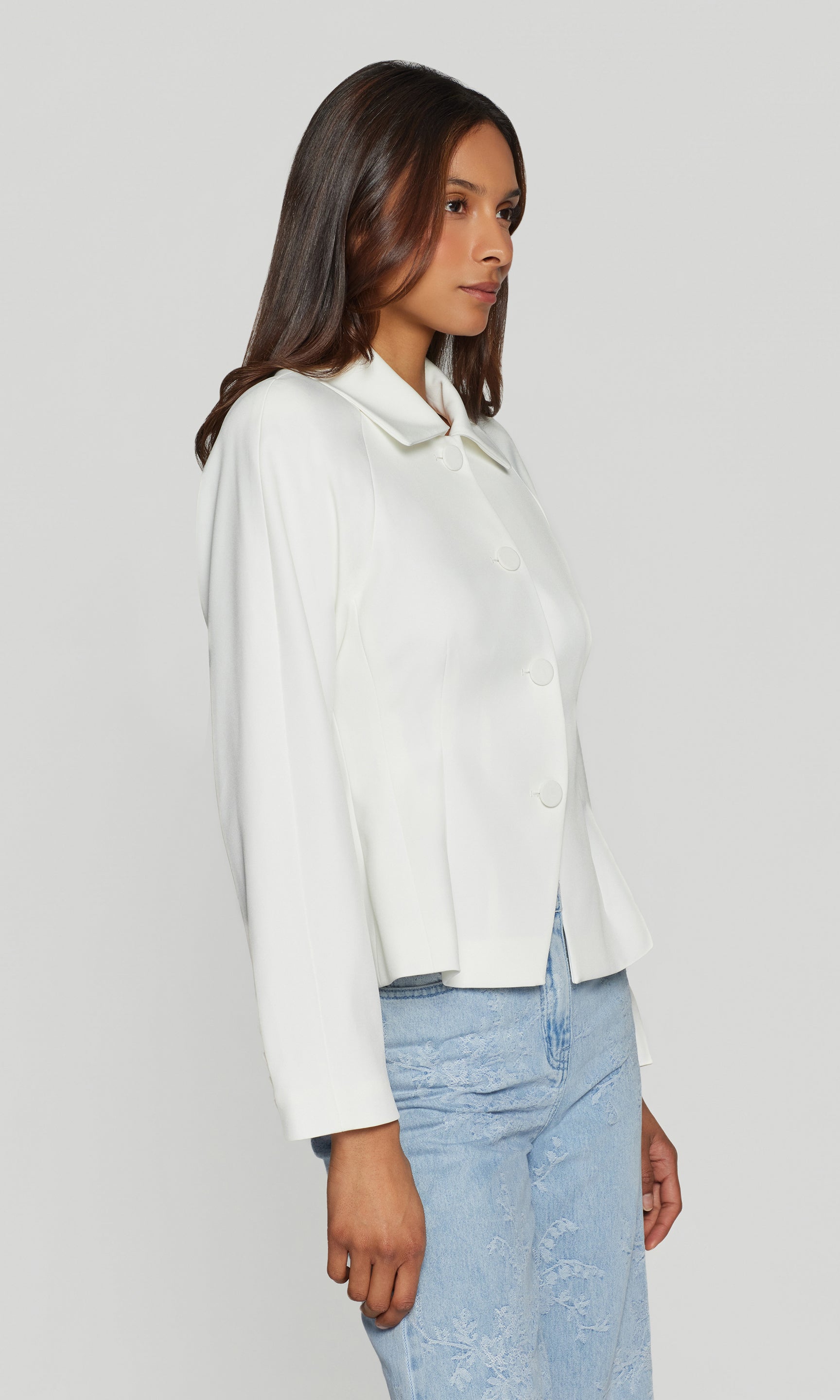 Side view of woman wearing a white jacket and light blue jeans against a plain background
