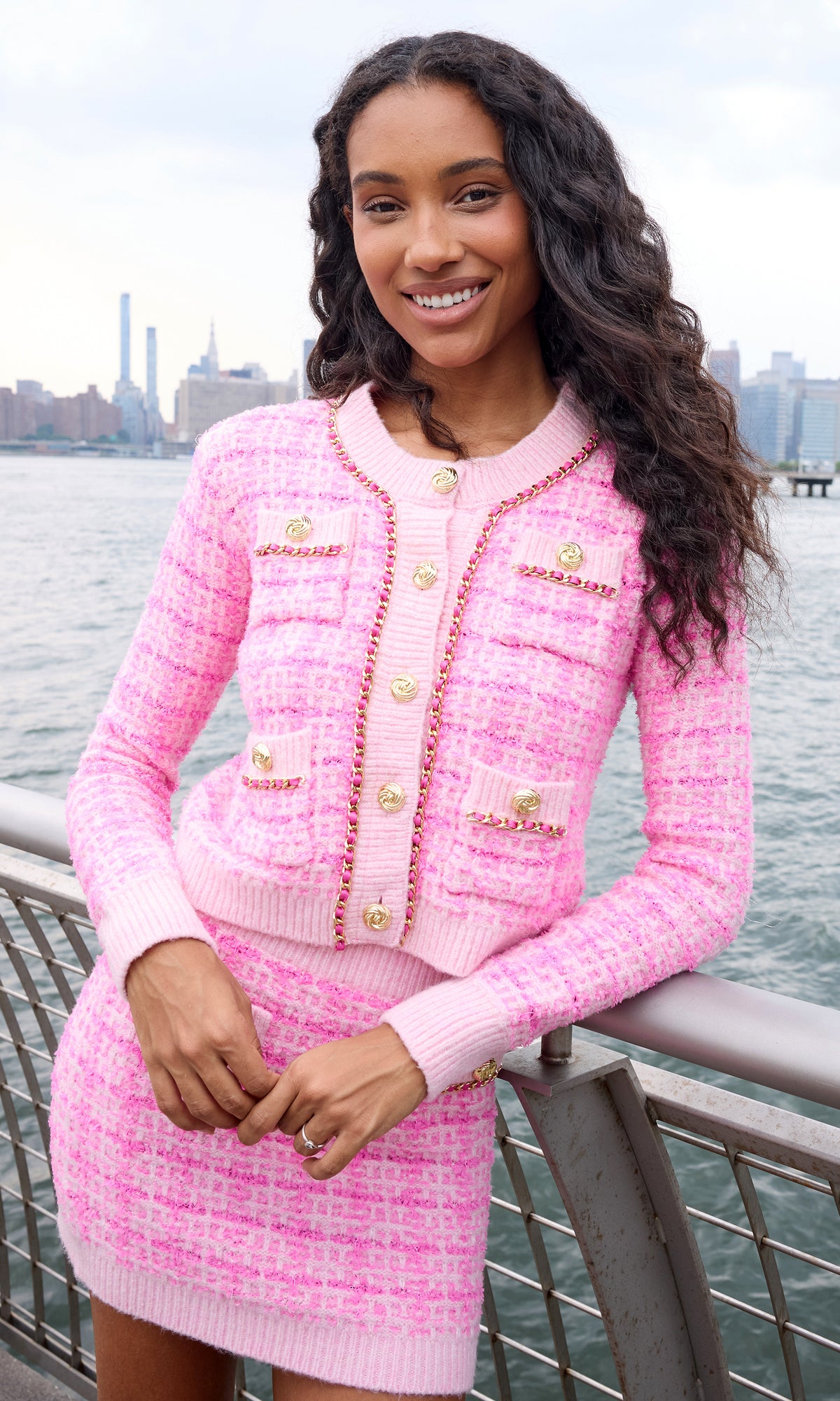 A woman wearing a pink tweed cardigan and matching skirt with gold-tone buttons and trim, standing outdoors near a waterfront railing with a city skyline in the background.
