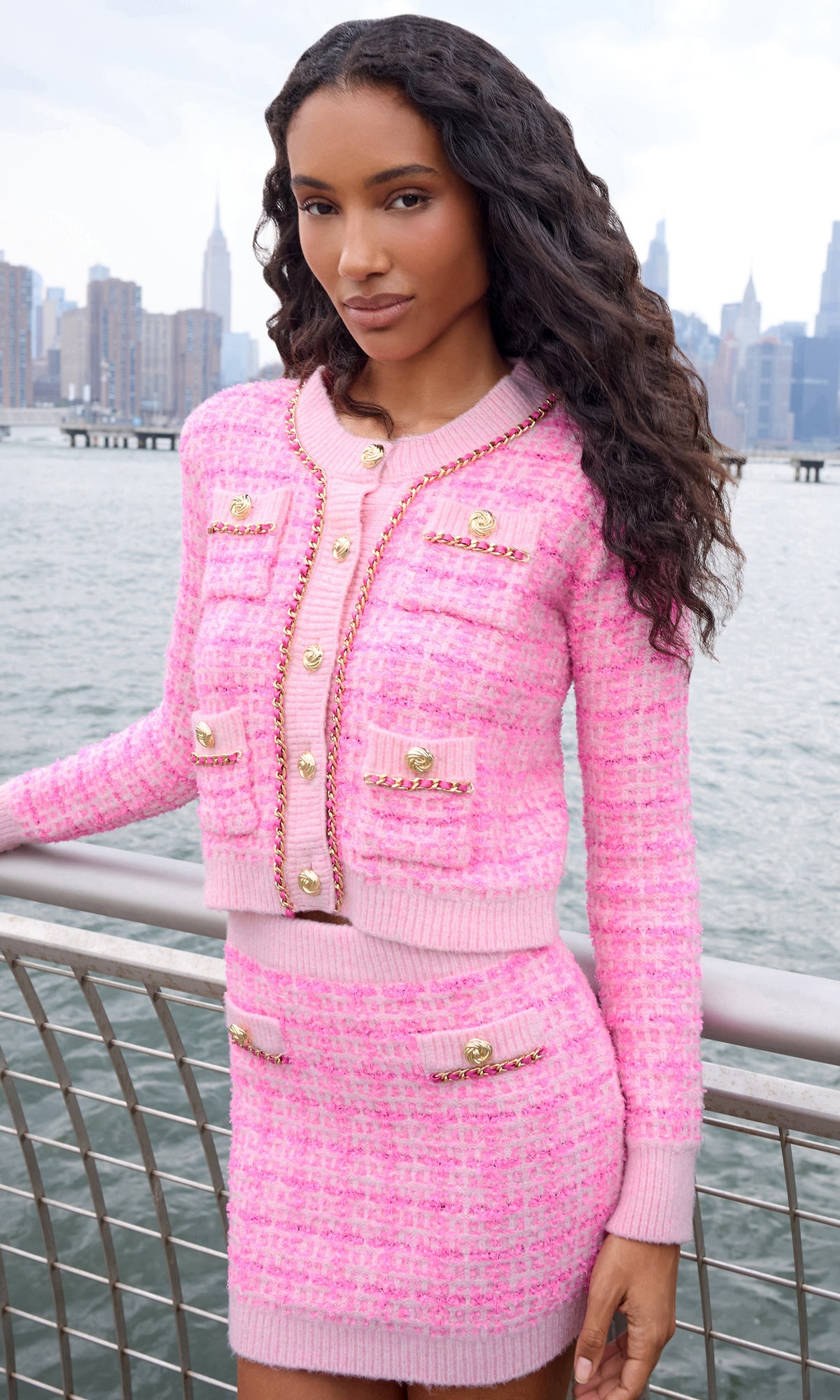 A woman wearing a pink tweed cardigan and matching mini skirt with gold-tone buttons and chain trim, posed outdoors with a city skyline and water in the background.