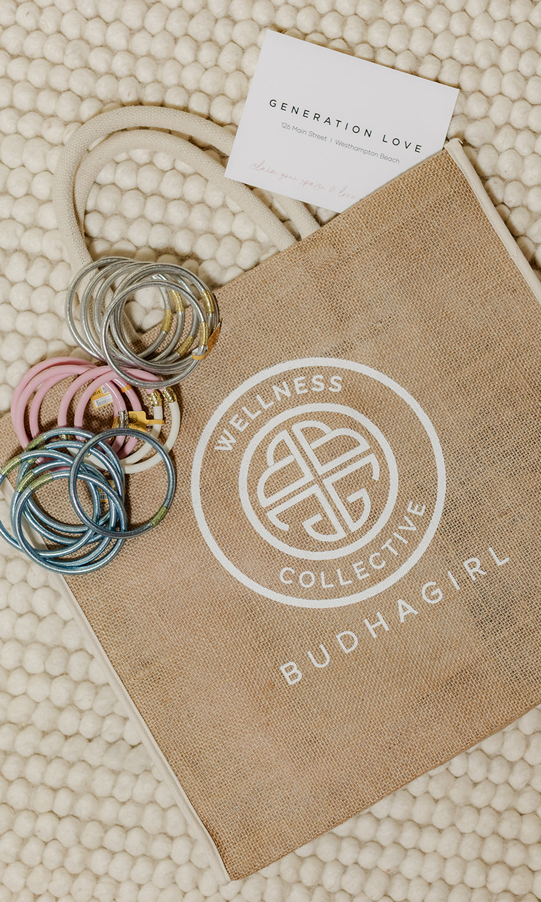 A  tote bag printed with 'WELLNESS COLLECTIVE BUDHAGIRL' in white, styled on a cream textured knit blanket with colorful metal bangles and a small card reading 'GENERATION LOVE'.