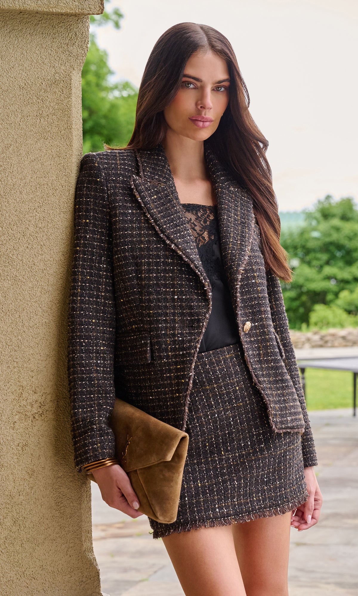 Model wearing a brown and black tweed blazer and matching mini skirt set, holding a brown suede clutch bag.