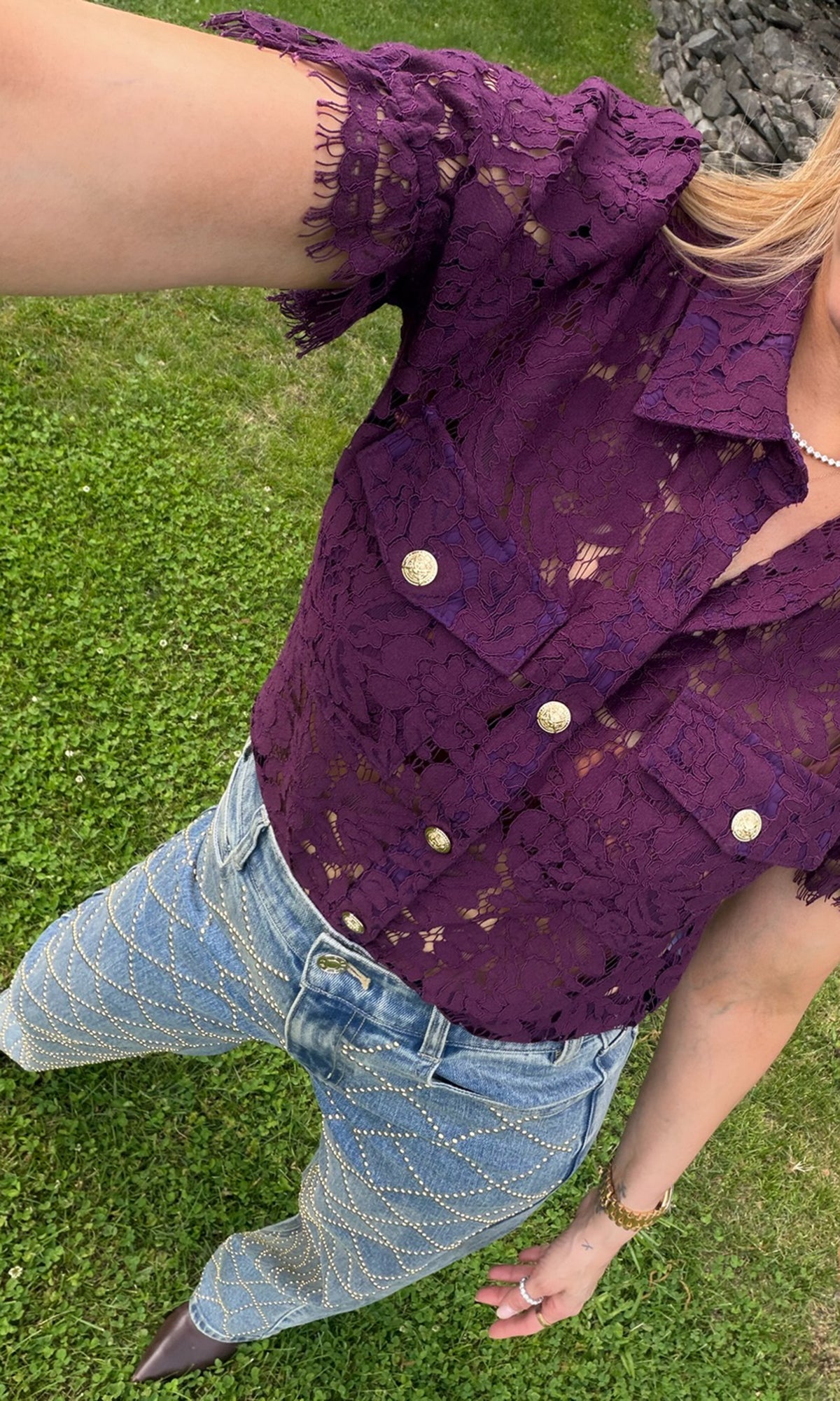 A person outdoors on grass wearing a short-sleeve purple lace button-front top with gold-toned buttons and light blue jeans with a gold chain pattern.