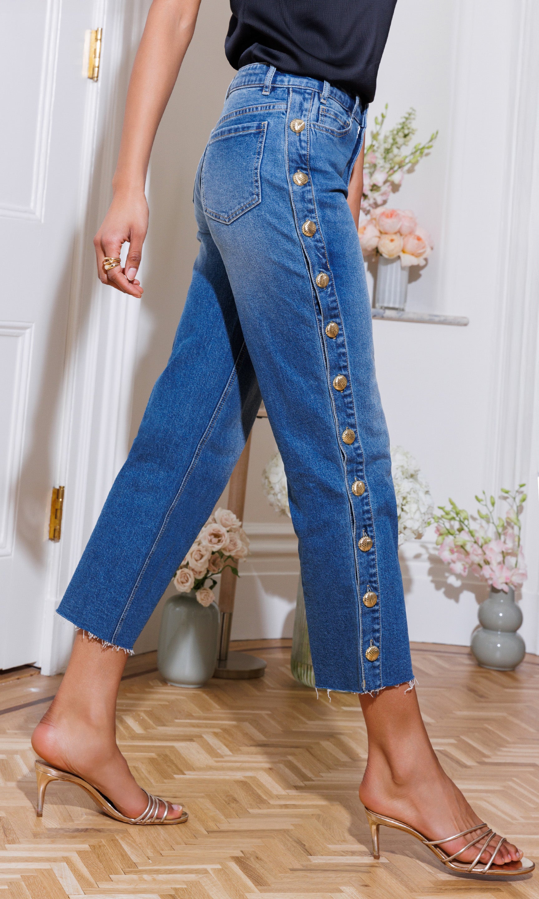 Person wearing high-waisted blue jeans with gold buttons in a room with flowers and a door.