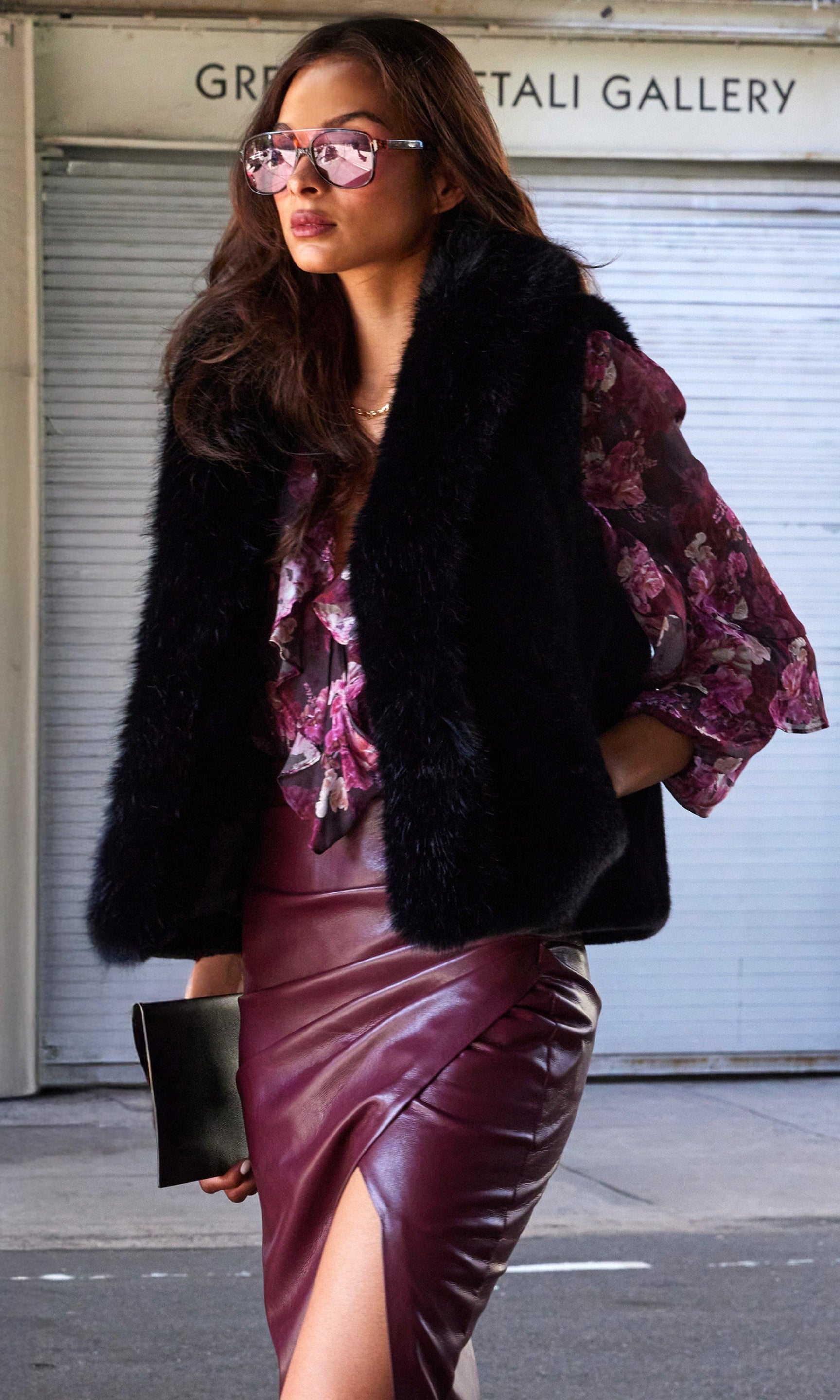 Woman wearing a black fur vest, floral blouse, and burgundy leather skirt in an urban setting.