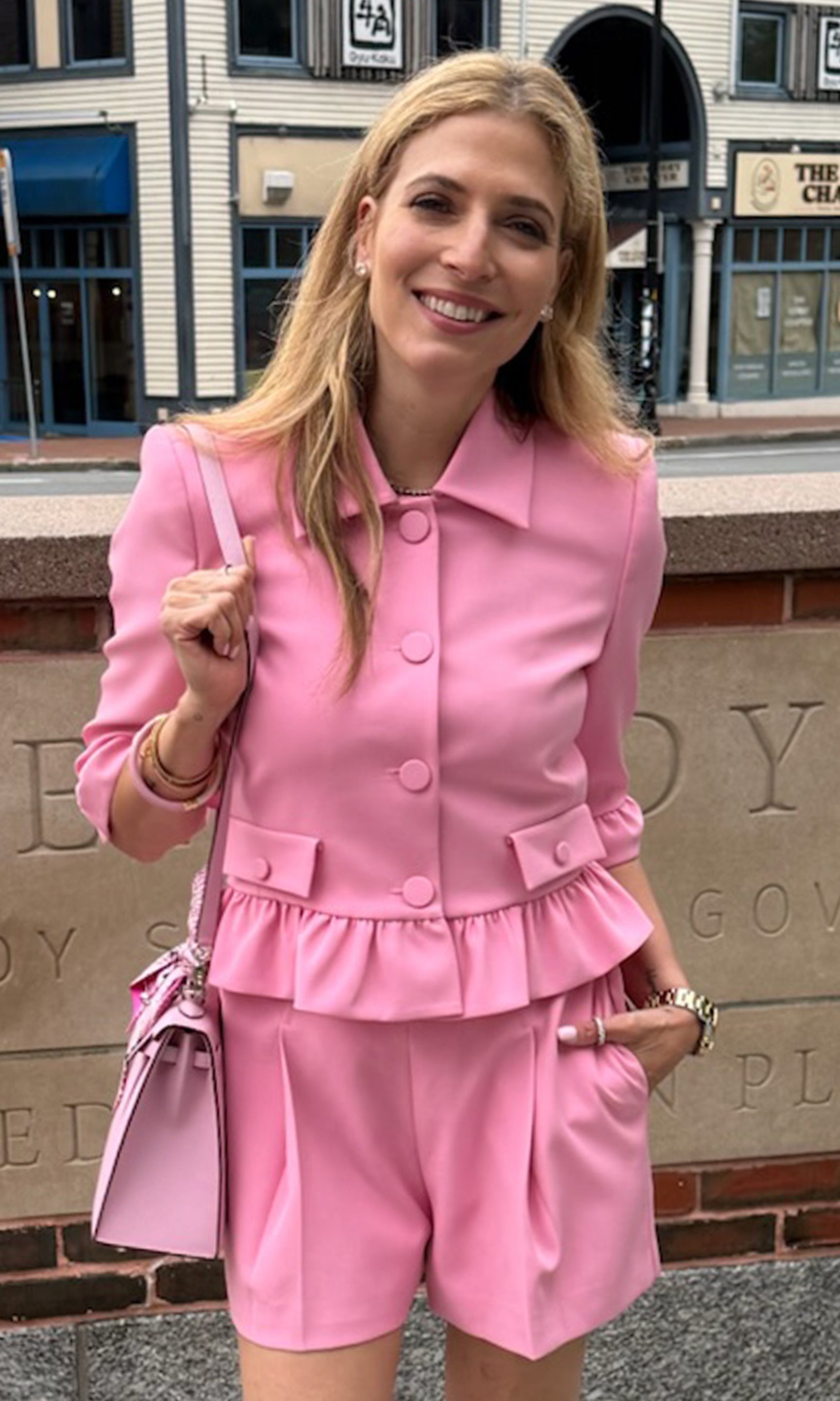 Founder Roni wearing a bright pink tailored jacket and matching shorts, standing outdoors in front of a stone wall.