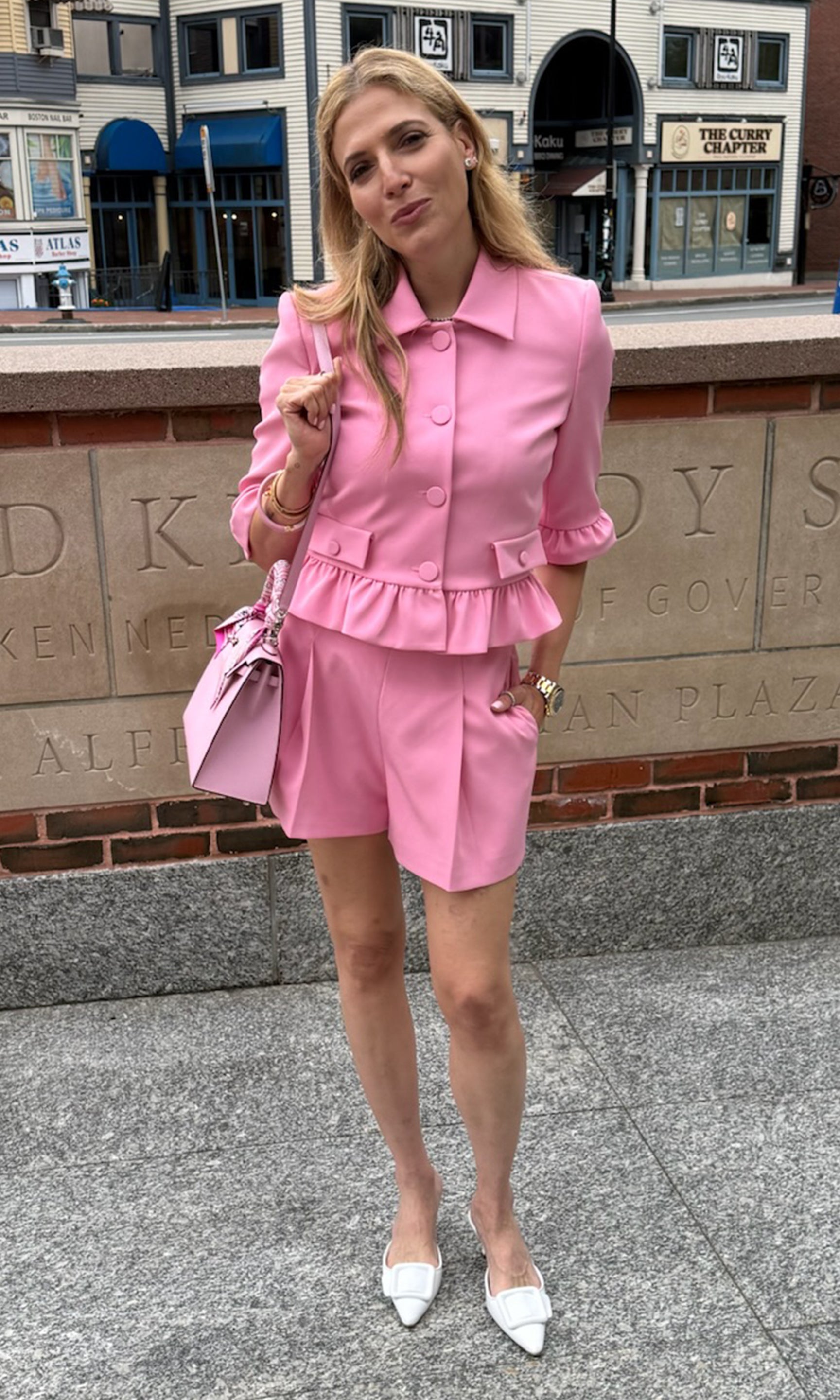 Founder Roni stands outdoors wearing a bright pink tailored matching set consisting of a short-sleeve collared jacket with a ruffled waist and matching shorts.