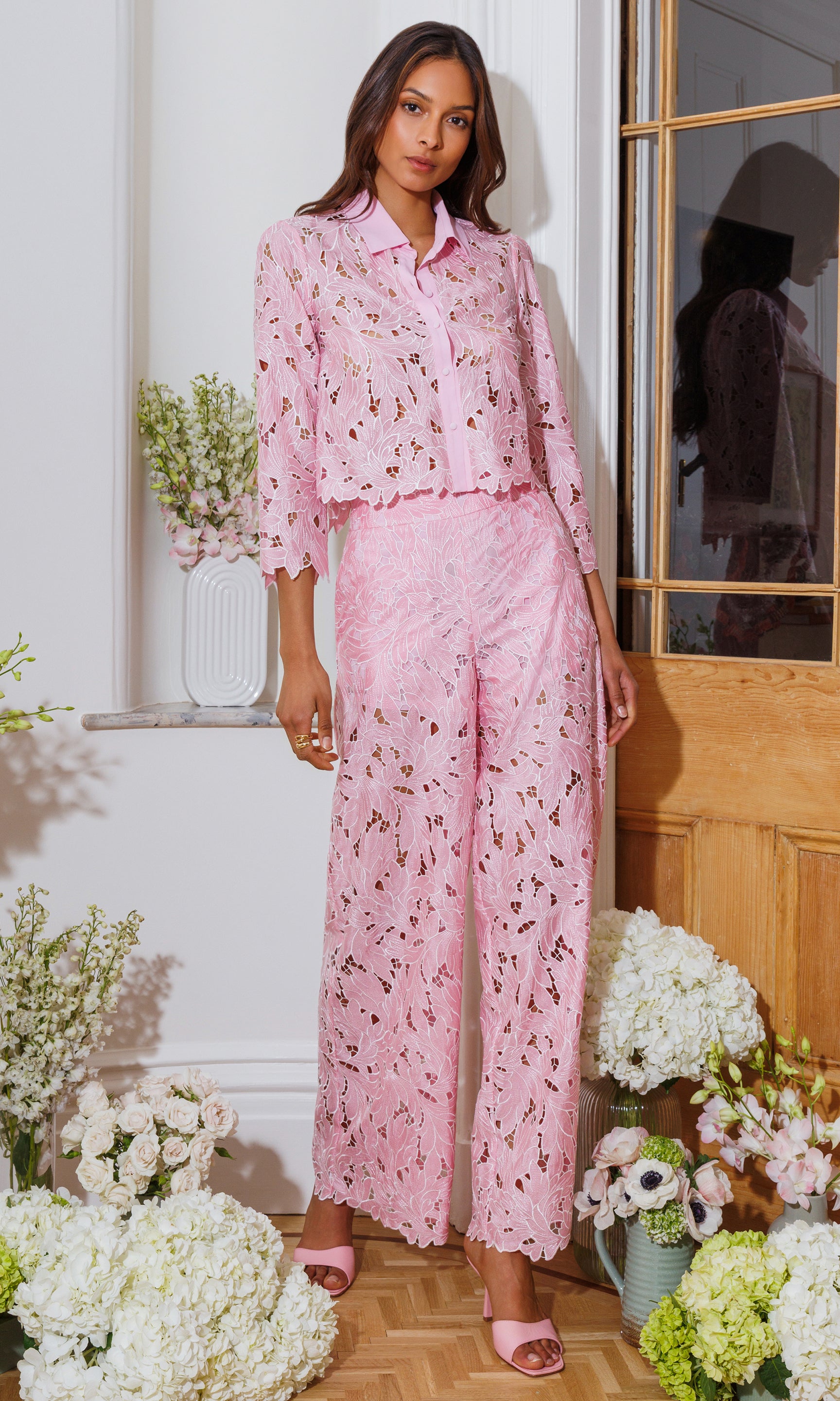 Woman wearing a pink lace outfit standing in a room with flowers and a wooden door.