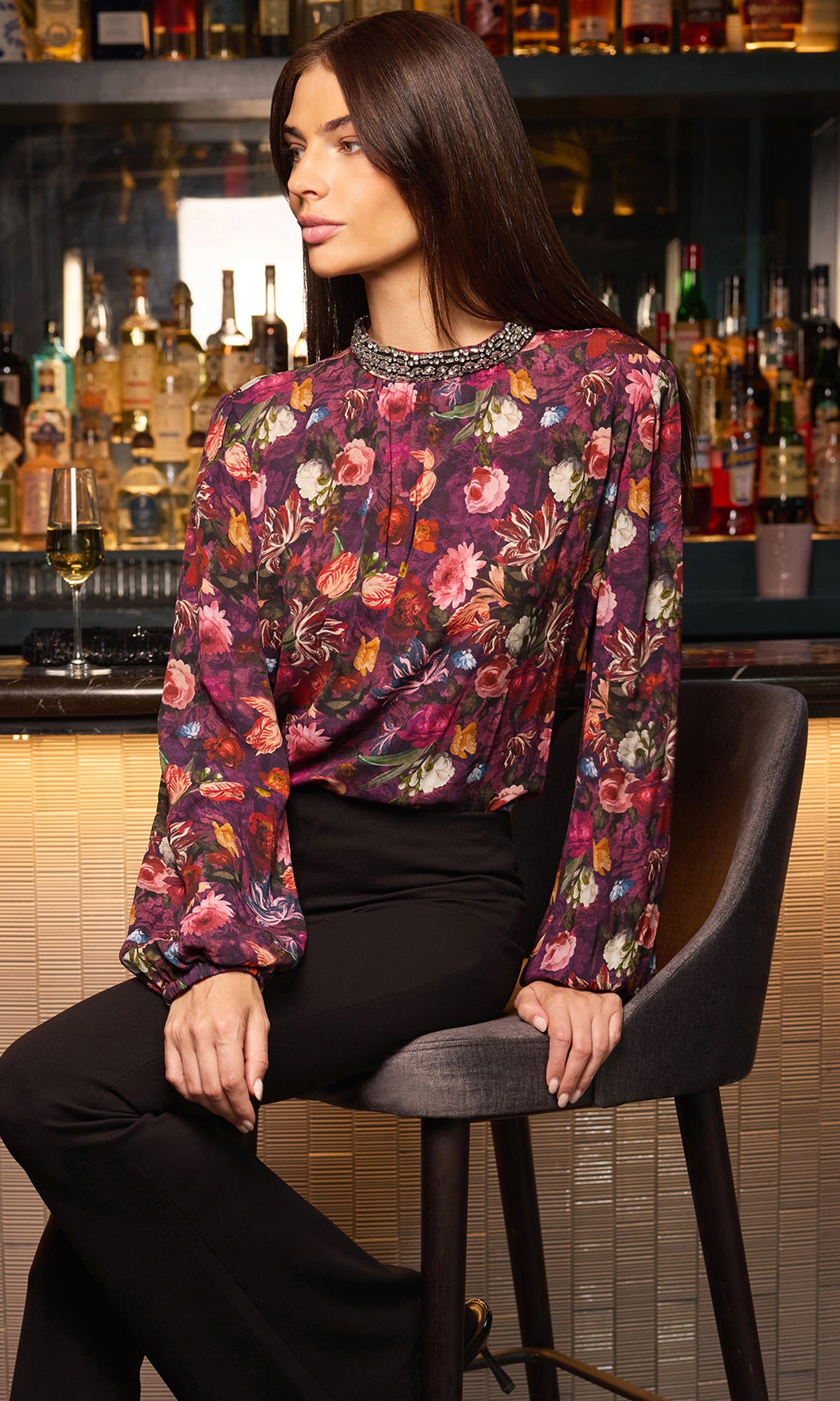 A woman seated on a bar stool wearing a long-sleeve floral blouse with a beaded collar and black pants, photographed in a bar setting with bottles and shelves in the background.