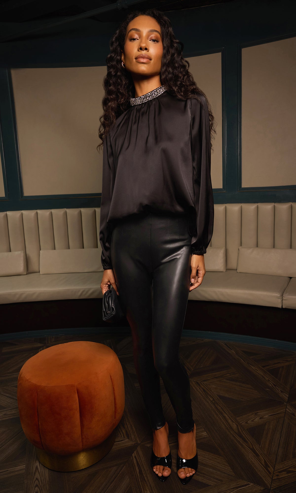 A woman stands in a lounge setting wearing a black satin long-sleeve blouse and black faux-leather leggings, with black high-heeled sandals and a jeweled choker.
