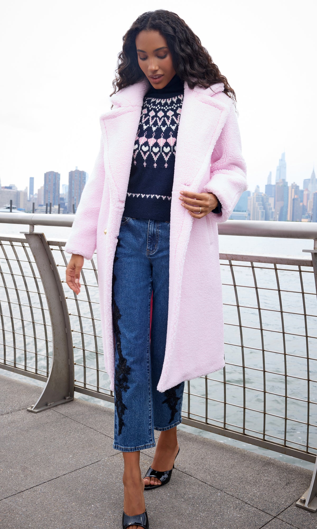 A woman stands outdoors near a waterfront railing wearing a light pink fuzzy coat over a navy patterned sweater, blue jeans, and black pointed flats, with a city skyline in the background.