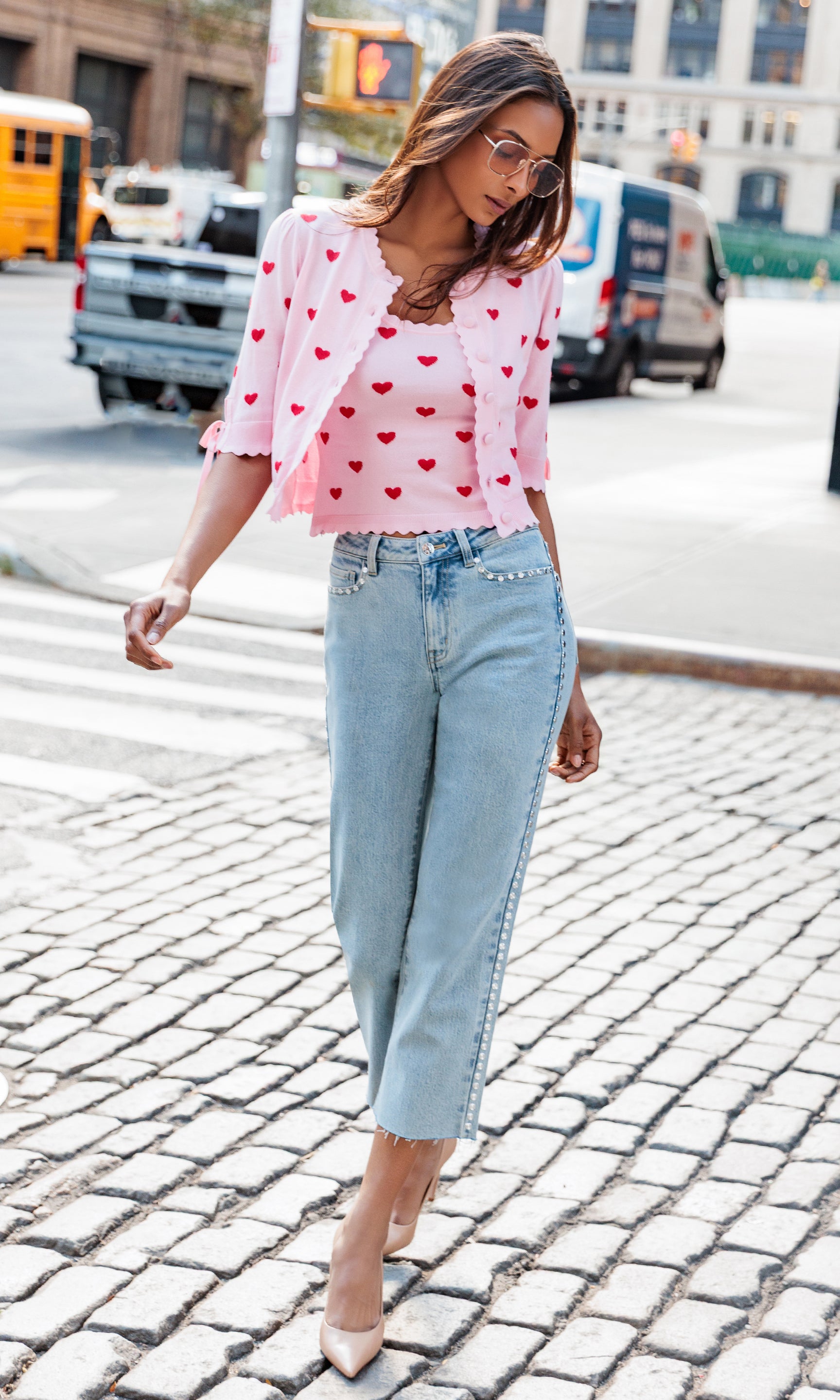 Woman wearing a pink heart-patterned blouse and light blue jeans on a city street.