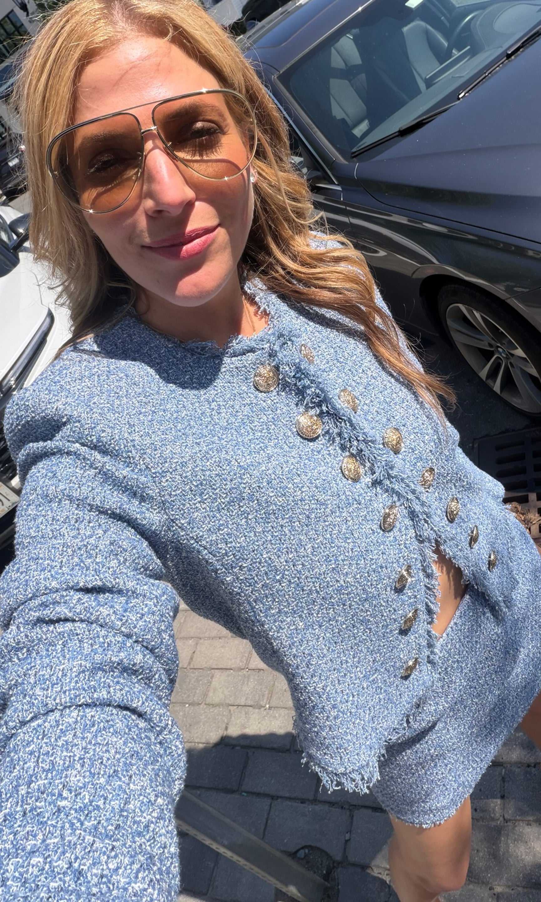 A woman takes a selfie wearing a light blue tweed jacket with gold buttons and matching shorts, standing outdoors next to a dark car.