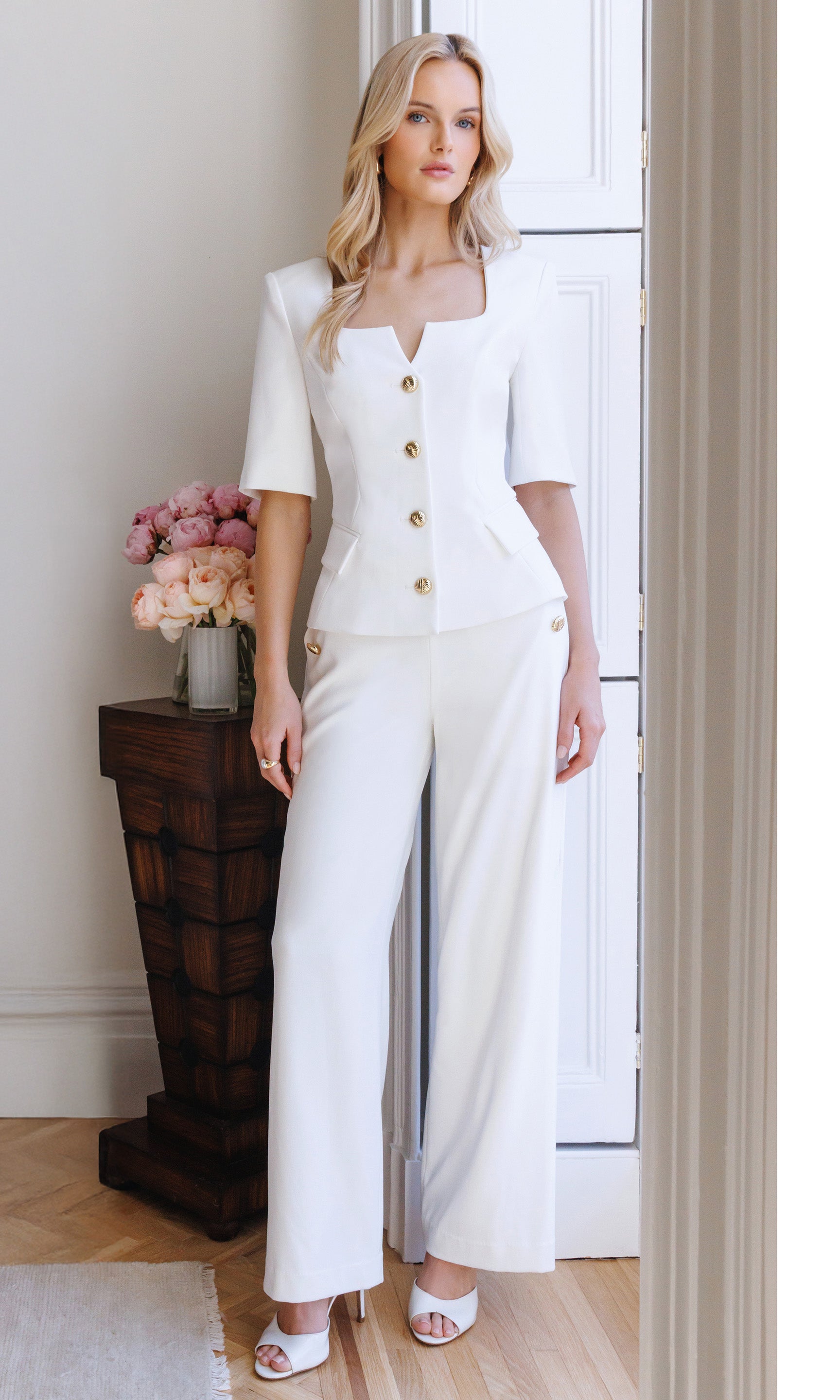 Woman wearing a white outfit with gold buttons in a room with a wooden side table and flowers.