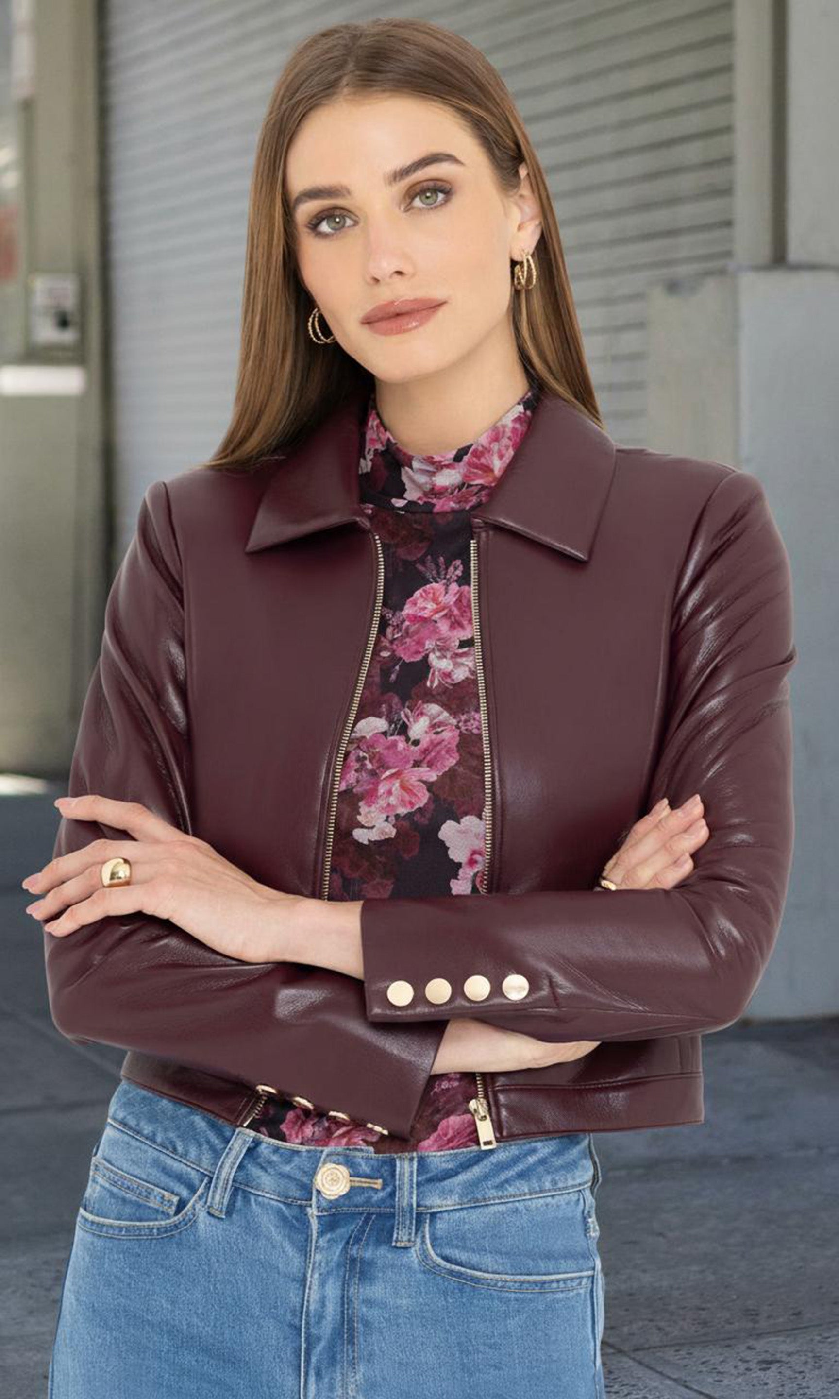 Woman wearing a brown leather jacket over a floral shirt with blue jeans, standing against a neutral background.