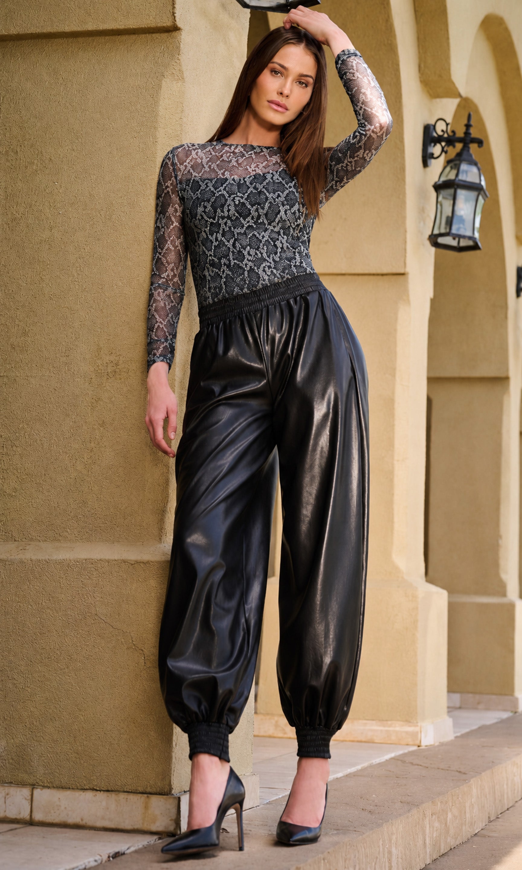 A woman models a black and gray patterned long-sleeve top paired with shiny black faux-leather harem pants, standing against a beige stucco wall.