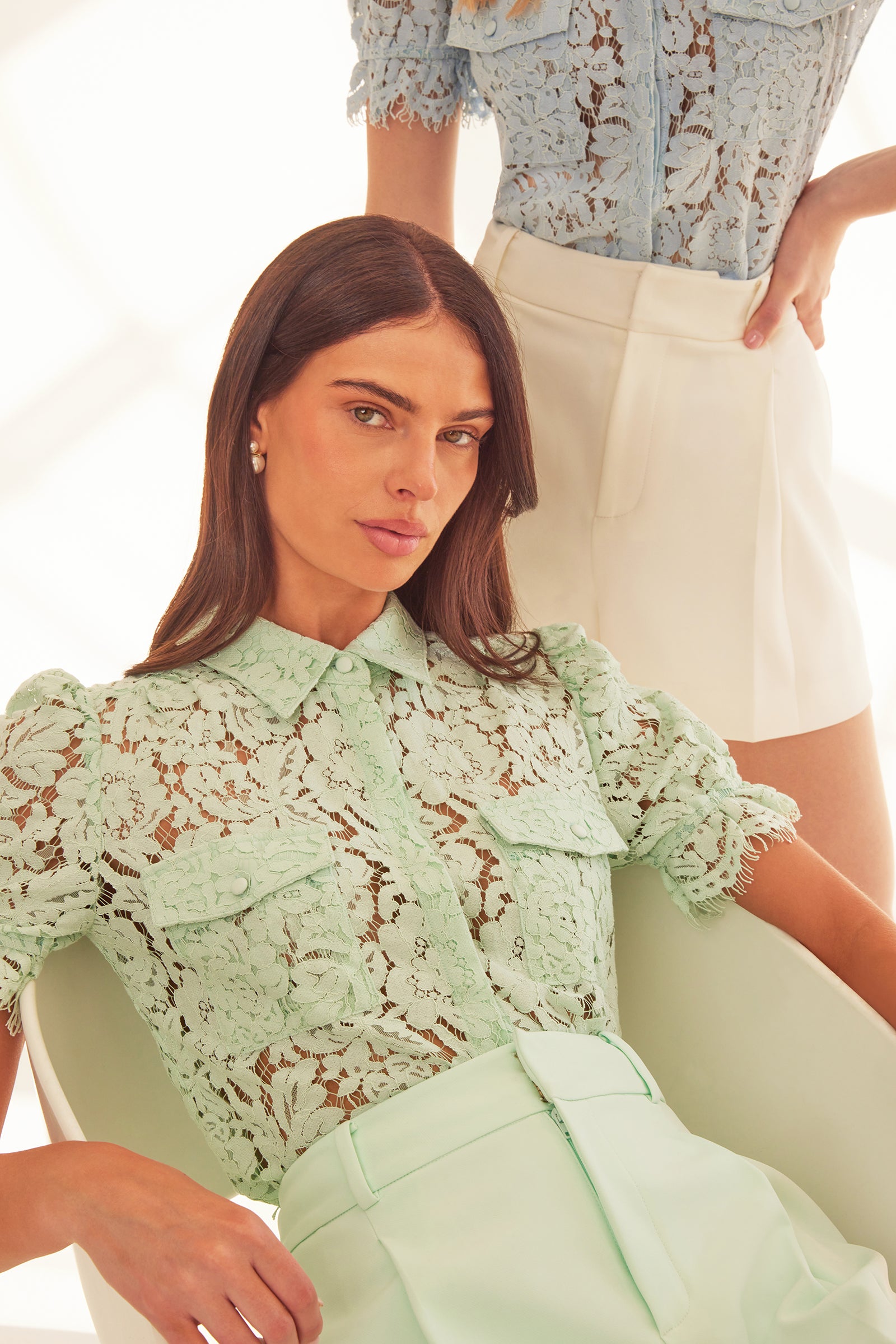model sitting on a chair while another model stands behind her. sitting in the front she is wearing a collar button shirt in the color seafoam with matching seafoam crepe shorts, in the back standing she is wearing the same shirt in light blue with white crepe shorts