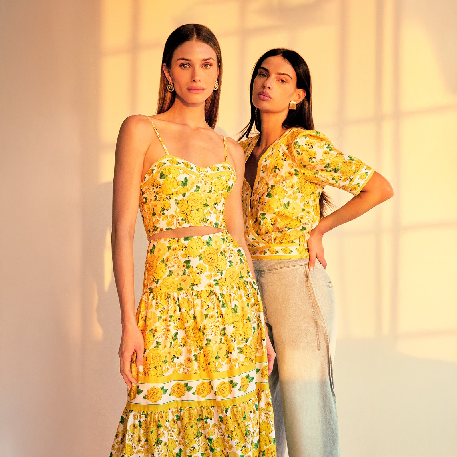 image of two models in a sunset-lit room both wearing styles from the same yellow floral pattern, on the right she is wearing the Rema Blouse with light blue jeans and on the left a bra cami top with a matching maxi skirt.