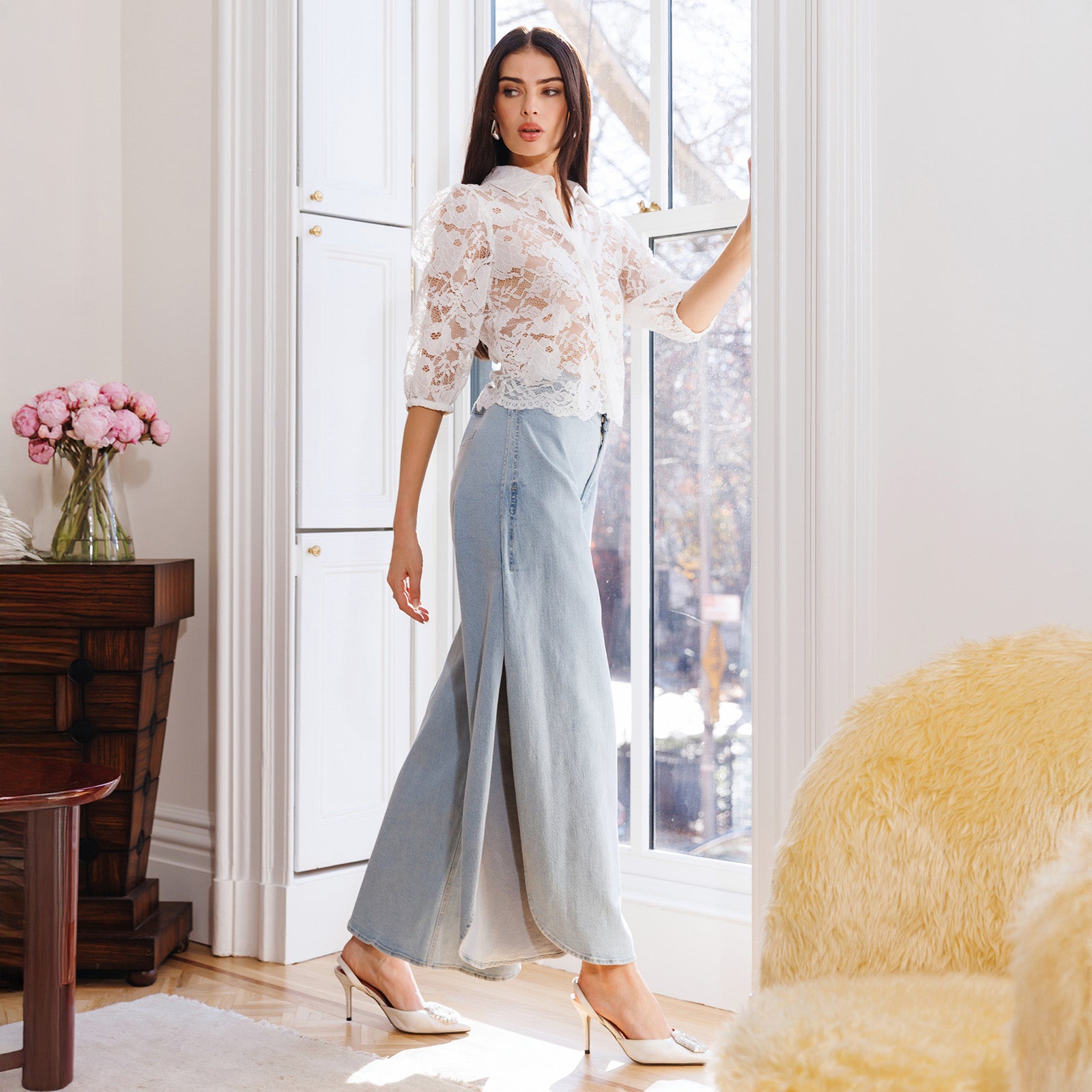 Woman in a white lace blouse and light blue pants standing in a bright room with large windows.