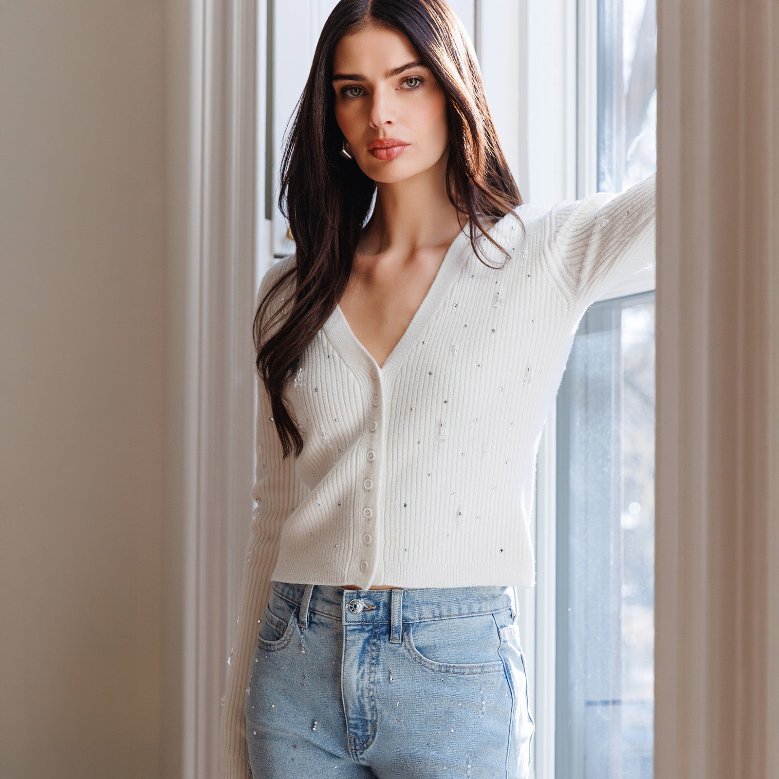 Woman wearing a white ribbed sweater and blue jeans standing indoors.