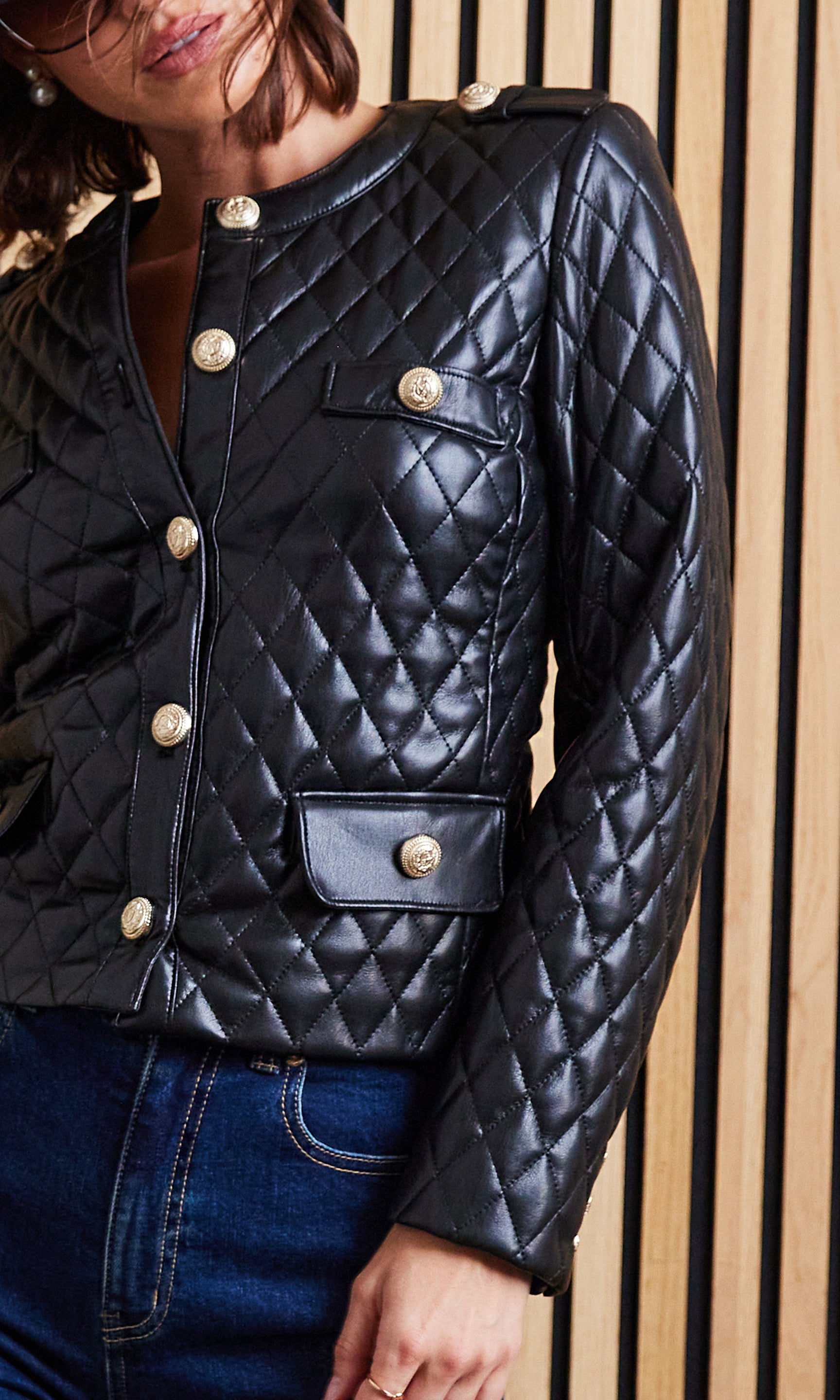 Black quilted jacket with gold buttons worn by a person against a wooden panel background