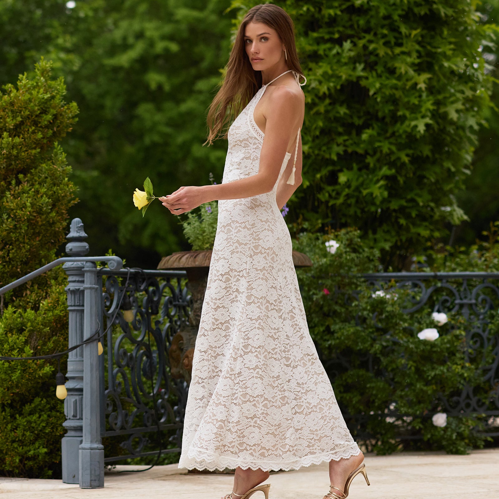 Woman in a white lace dress holding a yellow flower outdoors.