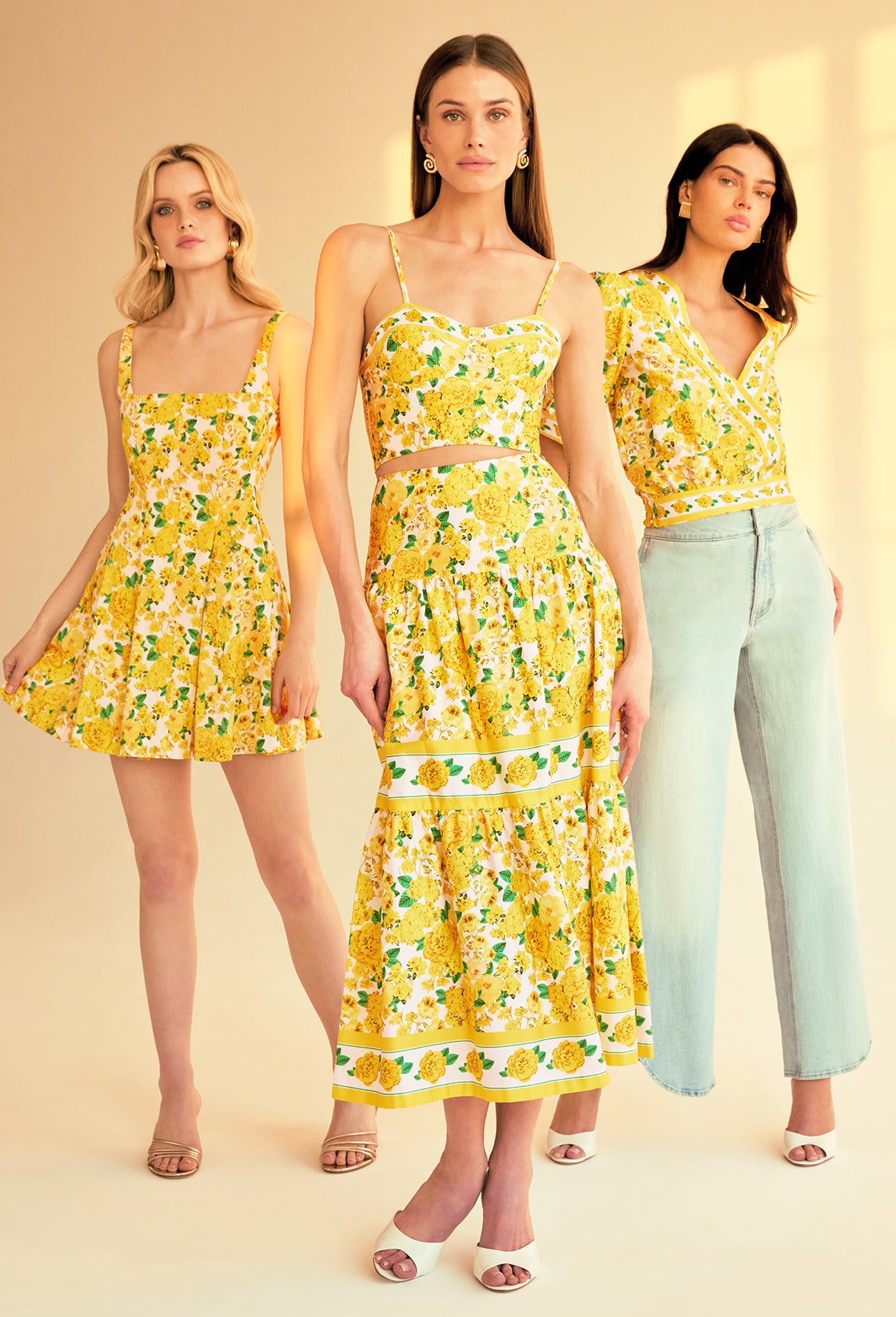 image of 3 models standing indoor a sunlit room all three wearing styles with the same yellow floral print, on the left she is wearing the Courtney mini dress, in the middle a bustier cami top with a matching skirt and on the right a v-neck blouse with blue jeans.
