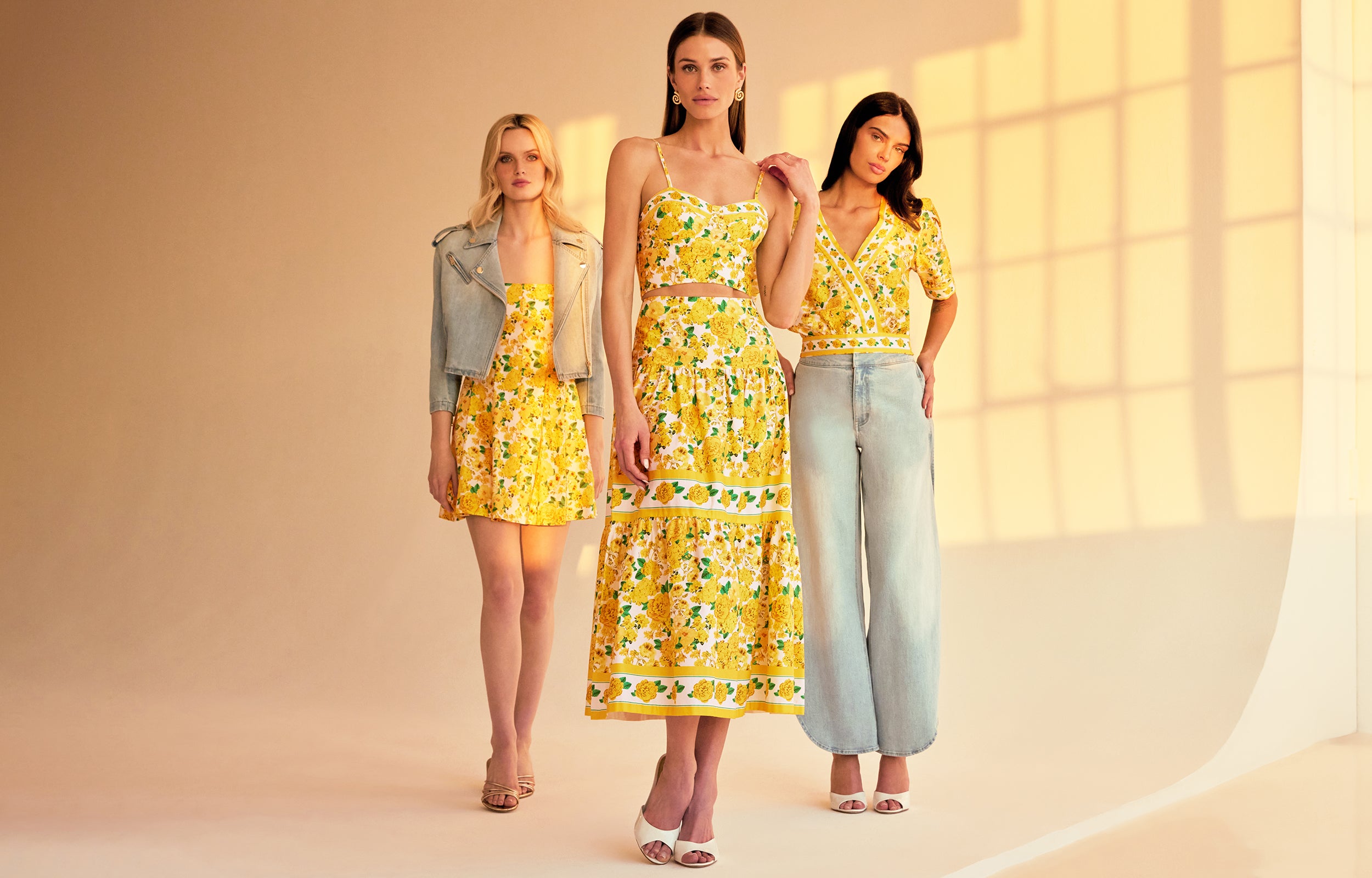 image of 3 models standing indoor a sunlit room all three wearing styles with the same yellow floral print, on the left she is wearing the Courtney mini dress, in the middle a bustier cami top with a matching skirt and on the right a v-neck blouse with blue jeans.