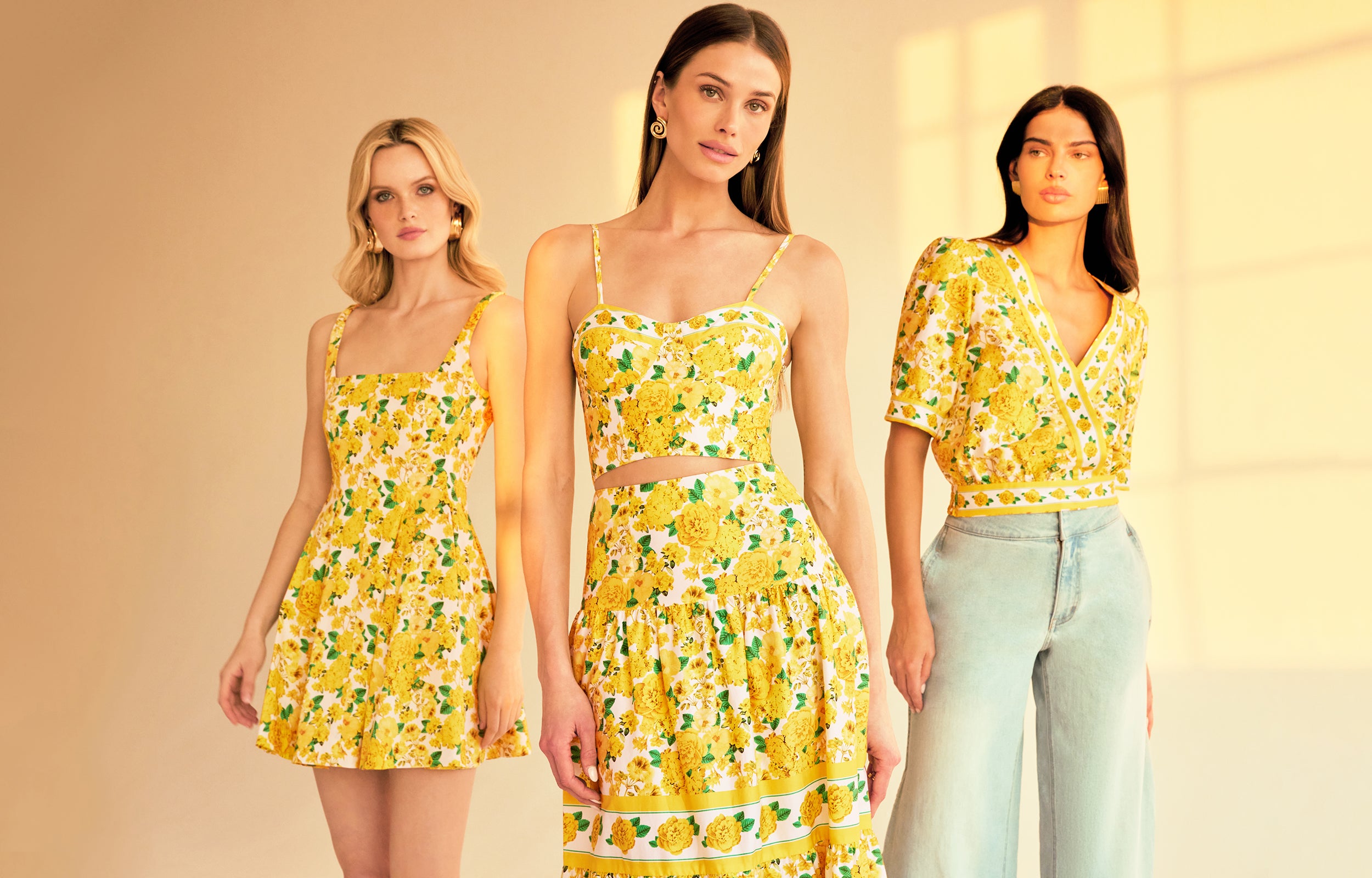 image of 3 models standing indoor a sunlit room all three wearing styles with the same yellow floral print, on the left she is wearing the Courtney mini dress, in the middle a bustier cami top with a matching skirt and on the right a v-neck blouse with blue jeans.