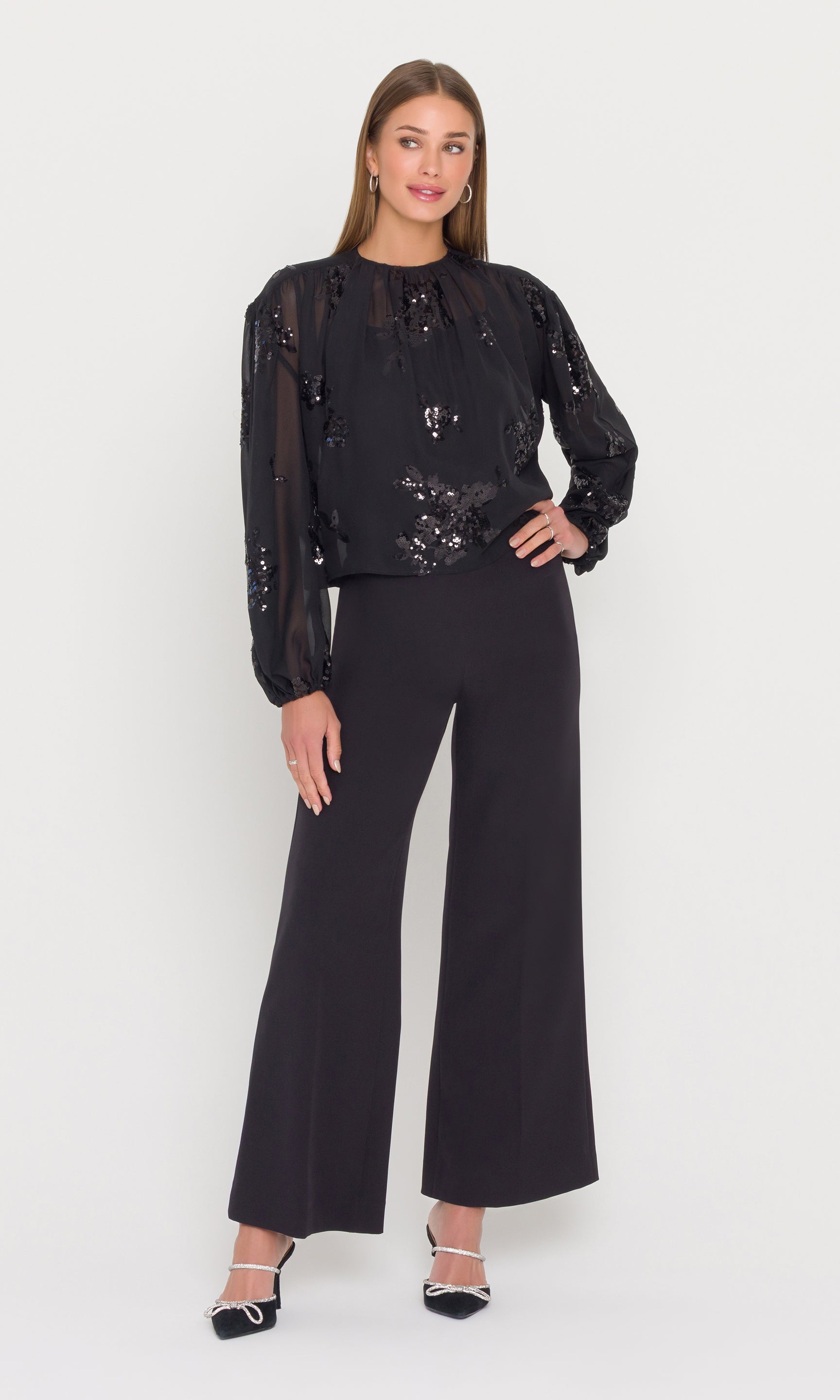 Woman wearing a black sheer top with sequins and black pants on a white background