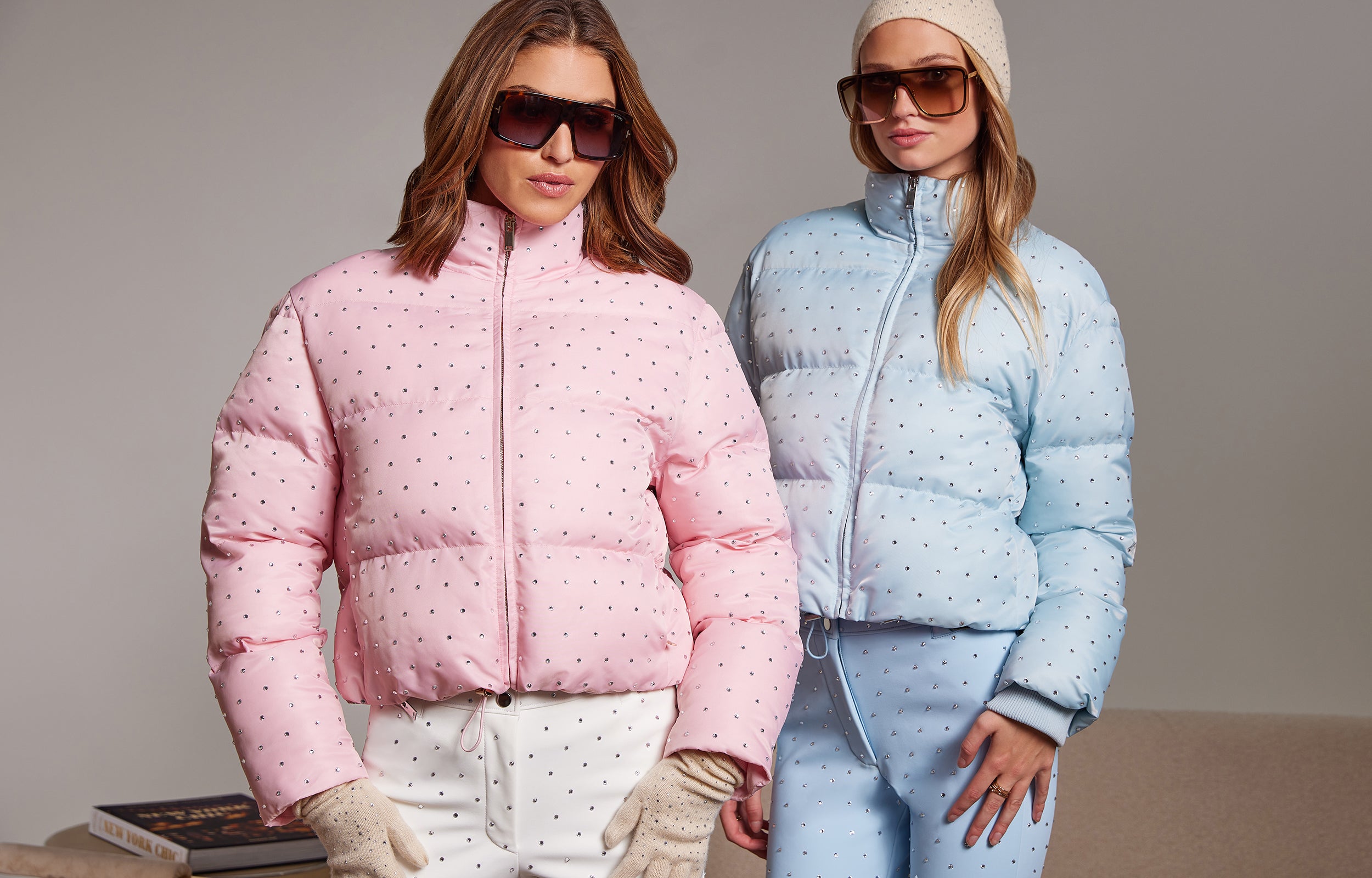 Two women wearing pink and blue puffer jackets with crystal embellishments on a neutral background.