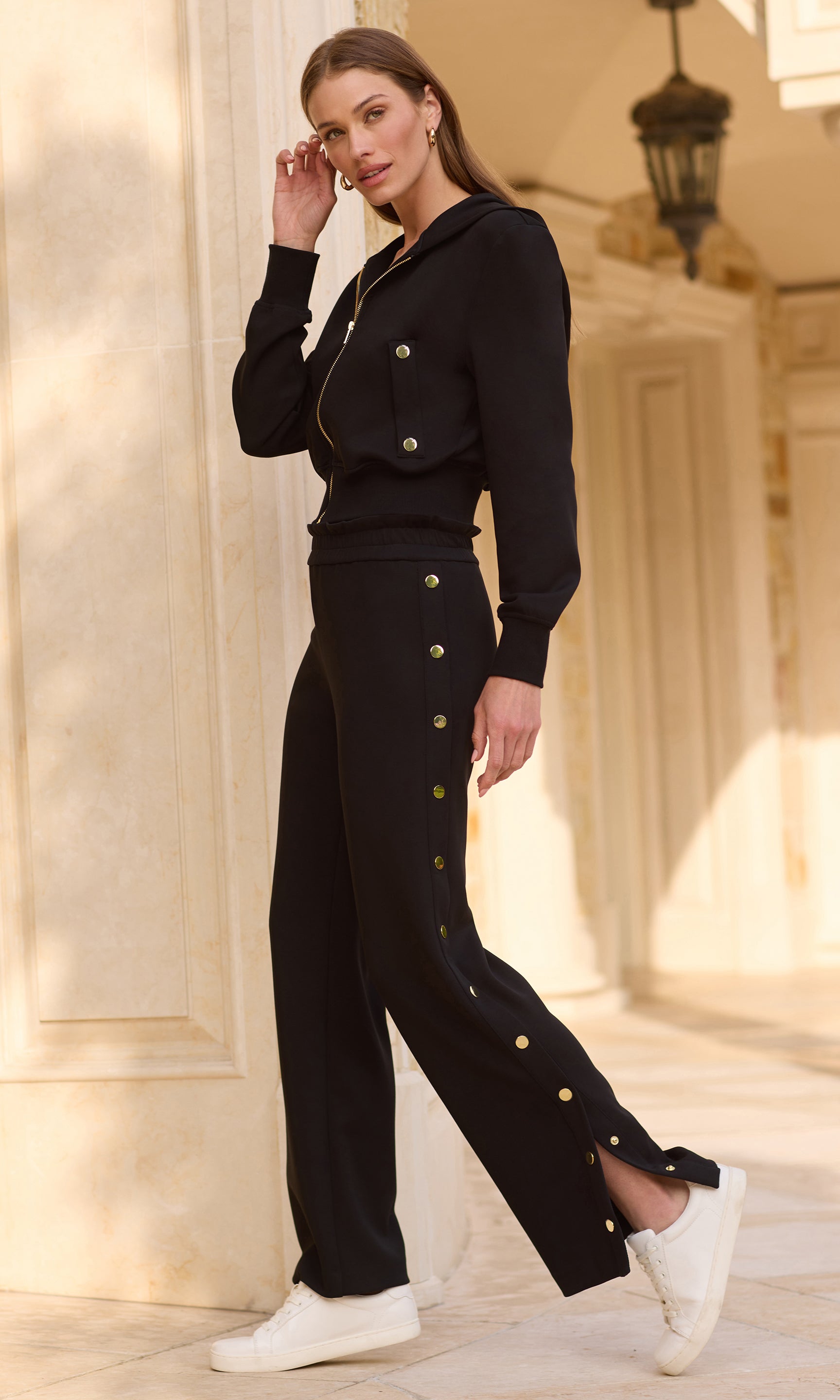 Side profile of model wearing a black sweatsuit with gold buttons on the jacket and side of the pant leg with white sneakers