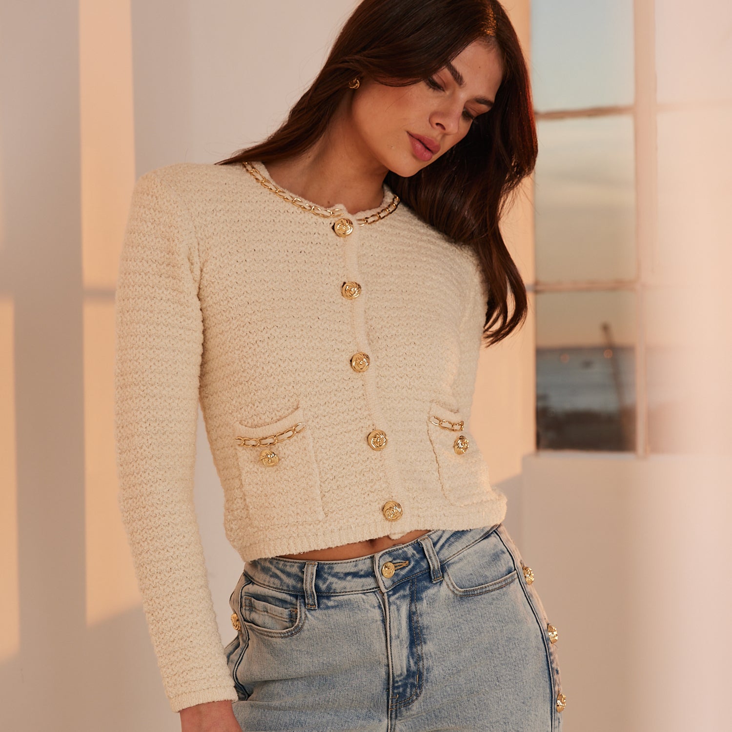Woman wearing a beige textured cardigan with gold buttons and blue jeans.