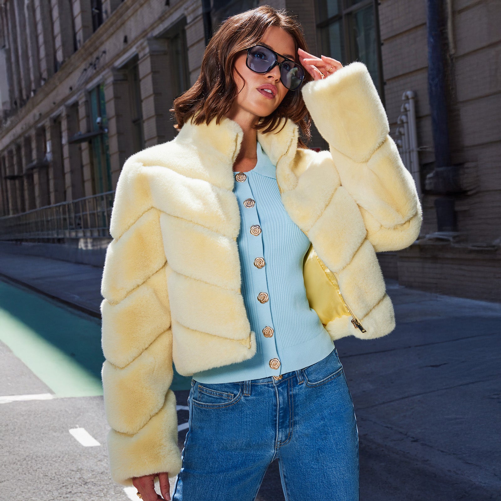 Woman wearing a light yellow faux fur coat, light blue sweater, and jeans on a city street.