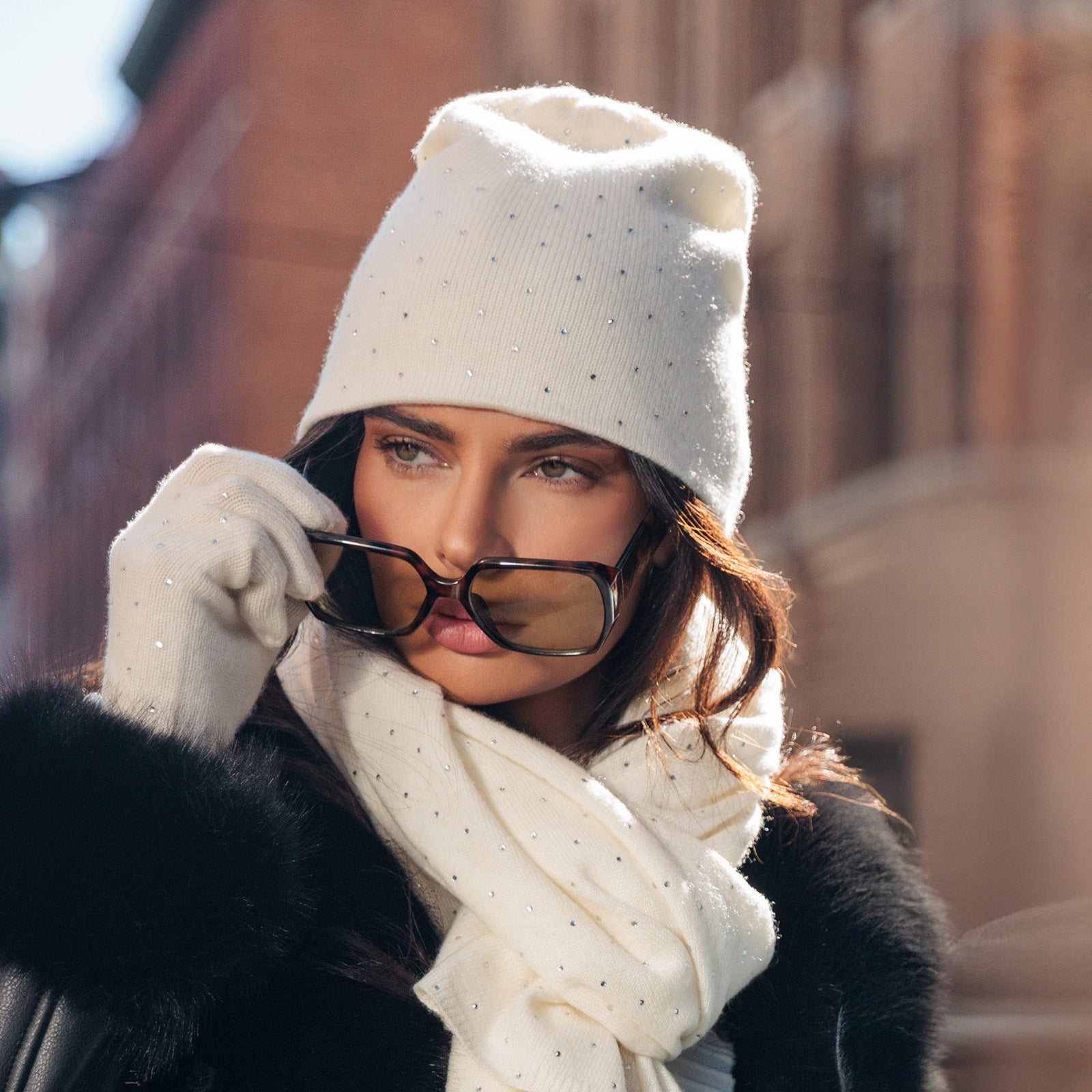 Woman wearing a white knit hat, scarf, and gloves with sunglasses outdoors.