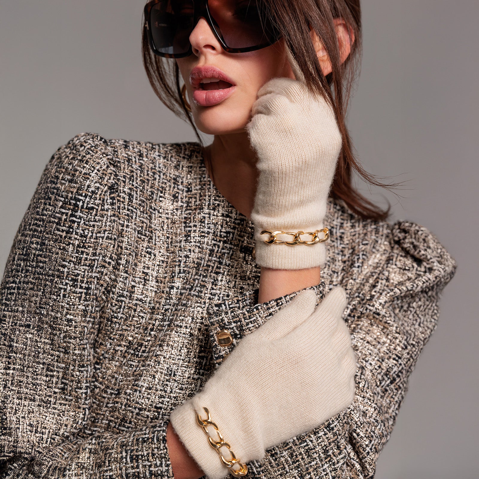 Woman wearing a textured coat, white gloves with gold chains, and sunglasses on a gray background