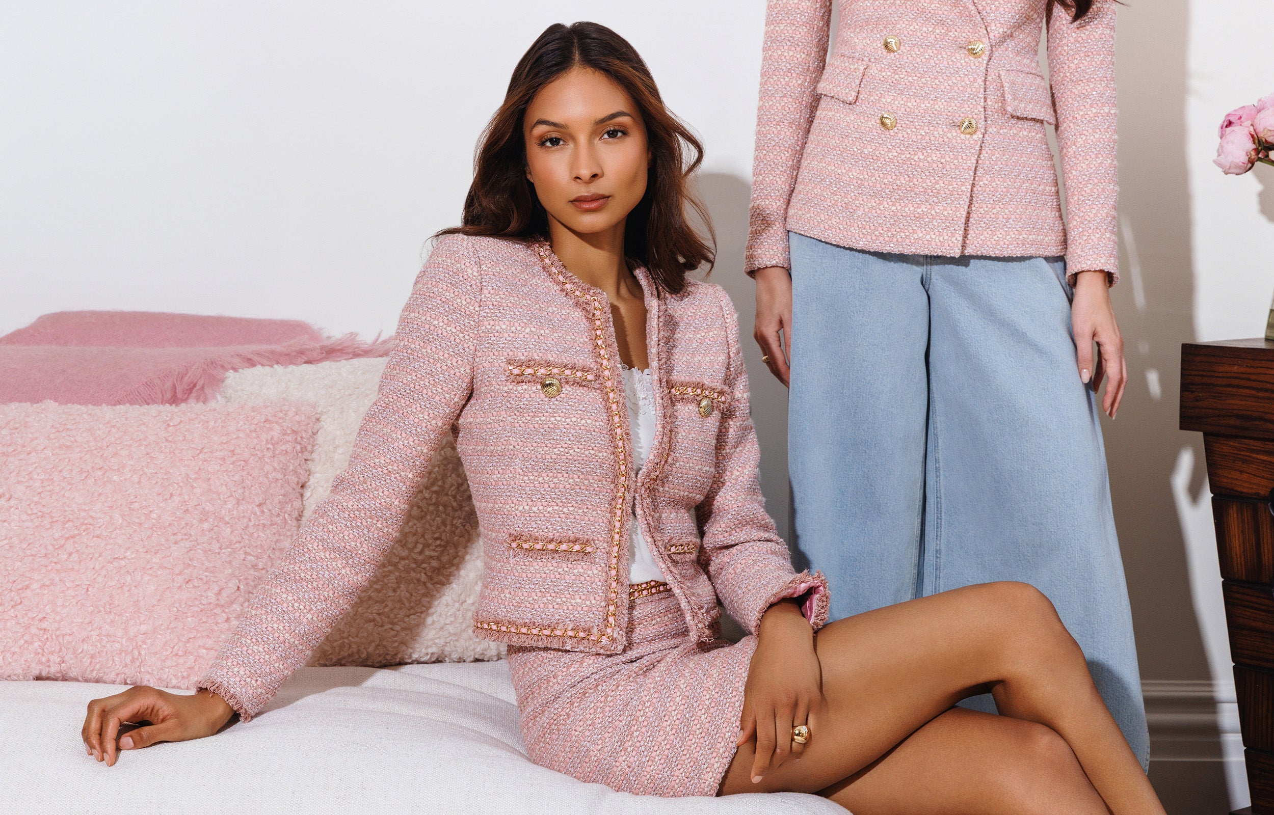 Woman in a pink tweed suit sitting on a bed with another person in the background.