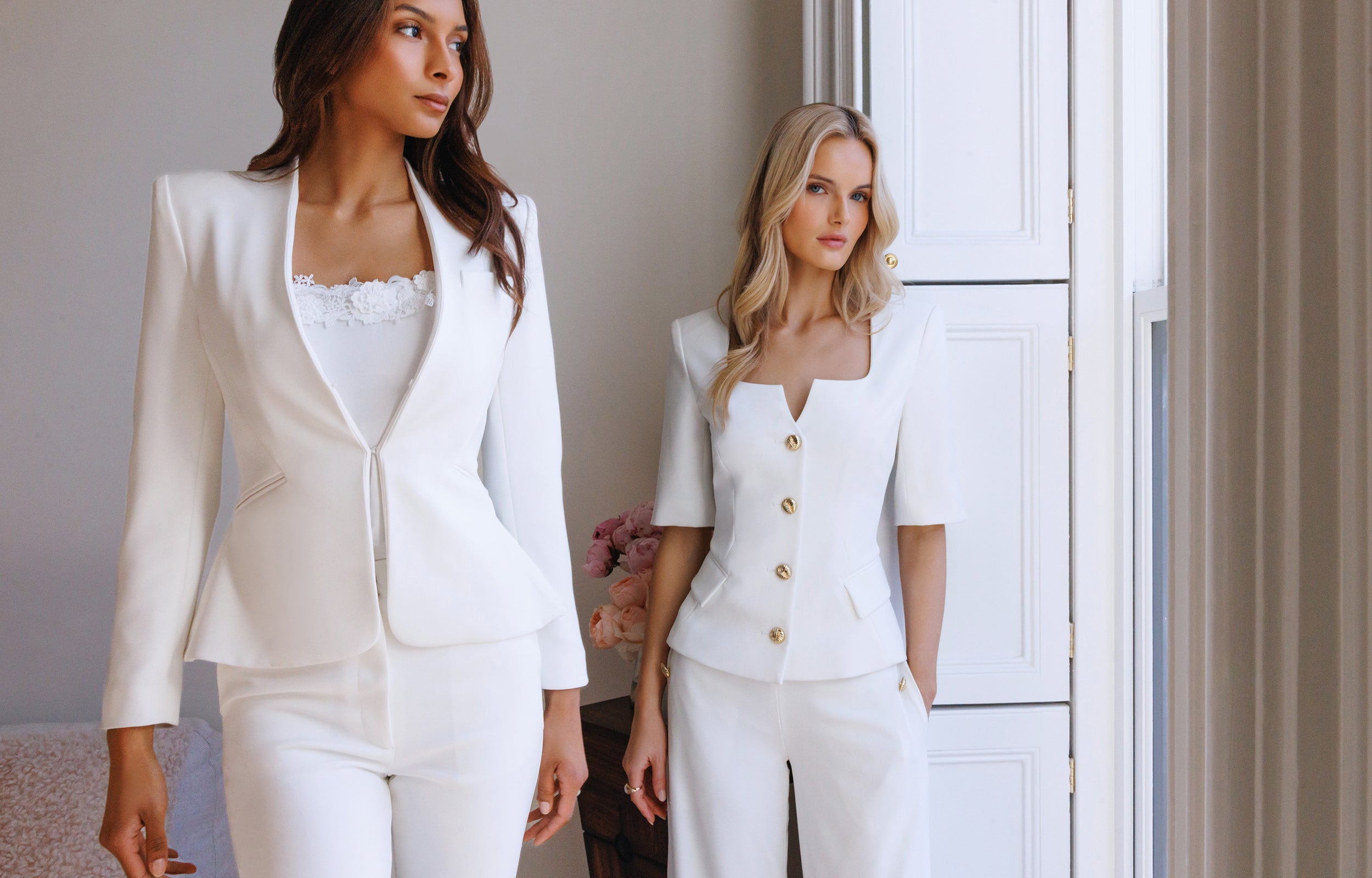 Two women wearing white suits standing in a room with neutral decor.