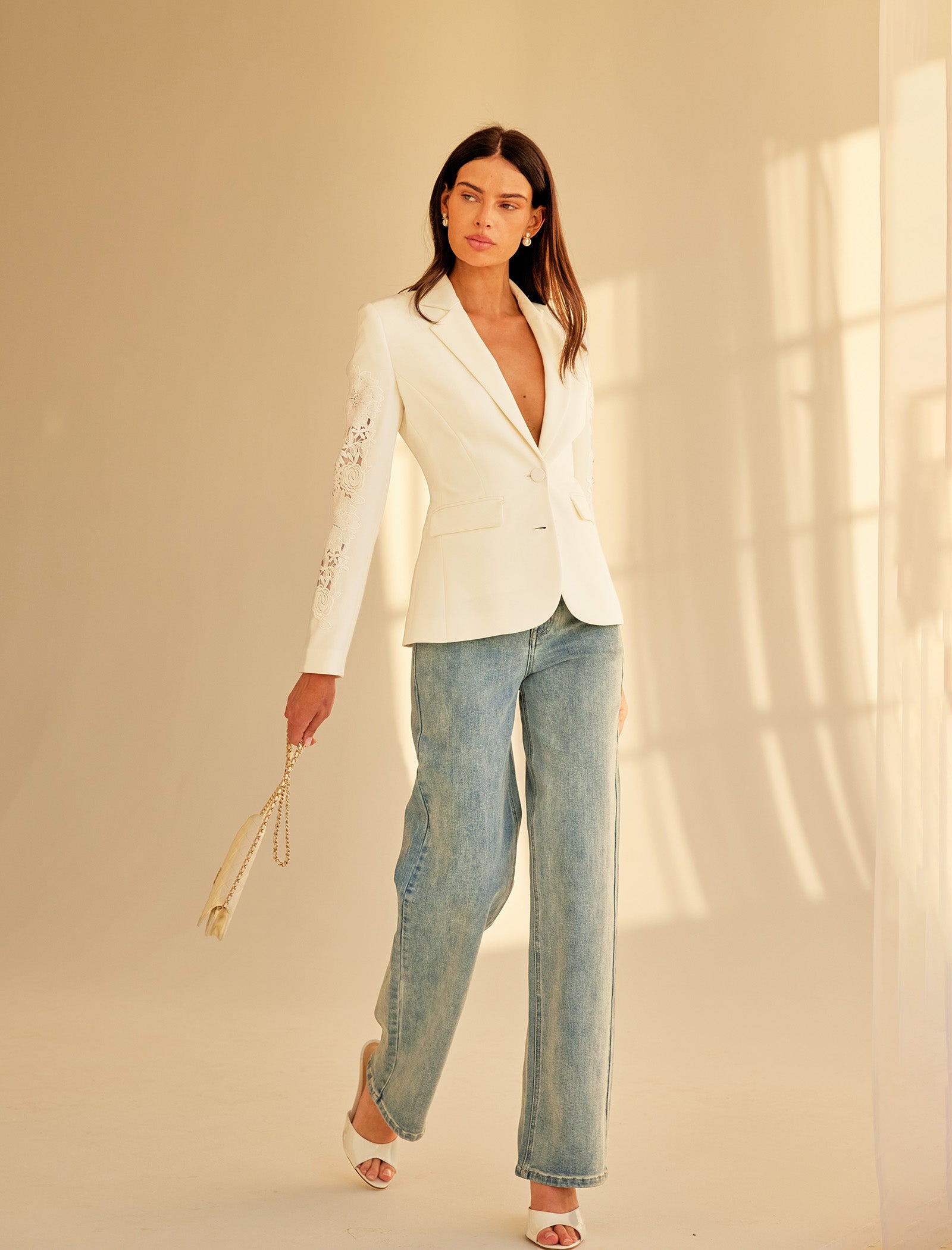 image of model wearing a white blazer with lace details on the sleeve paired with jeans holding a small white purse and wearing white heels