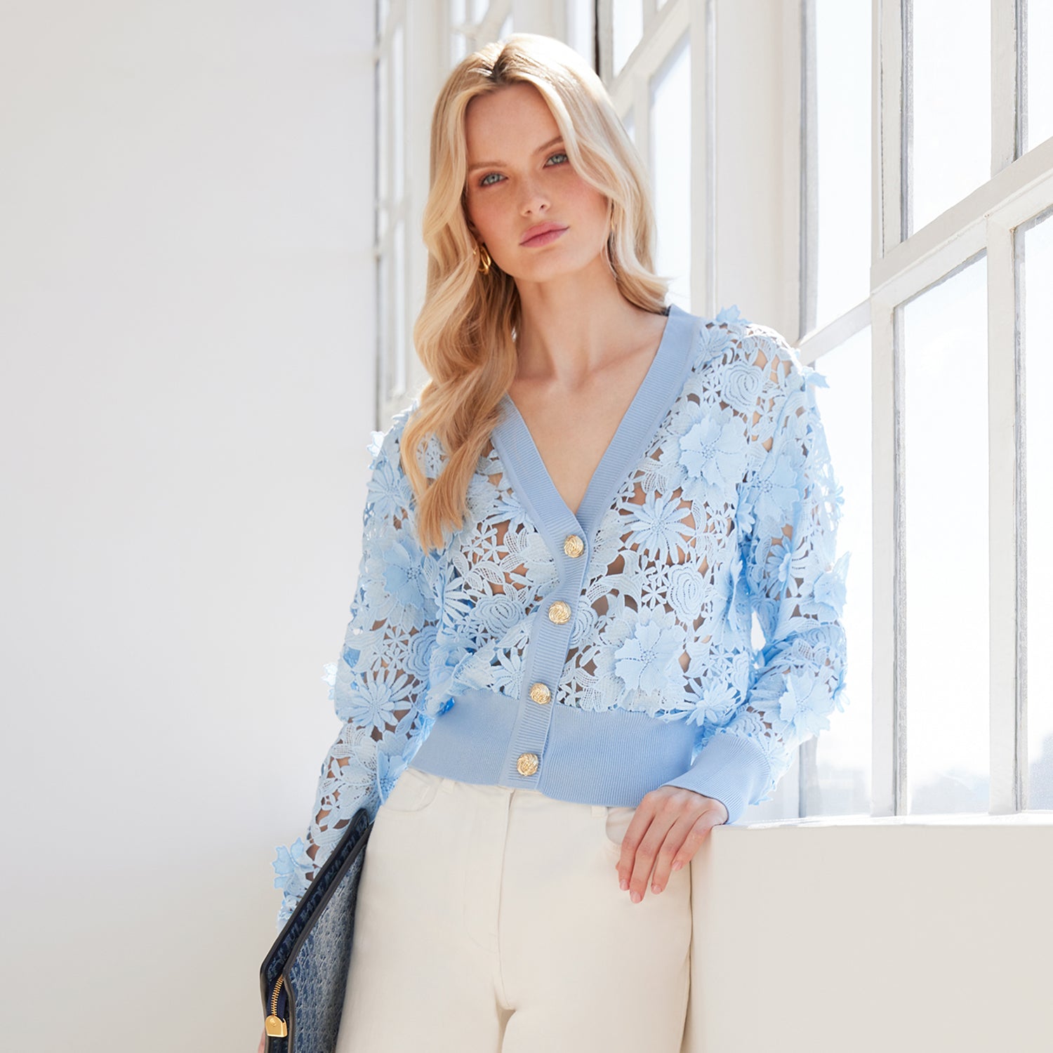 image of model posing in front of a window, wearing the Harlow cardigan in blue, paired with white jeans and holding a navy clutch purse