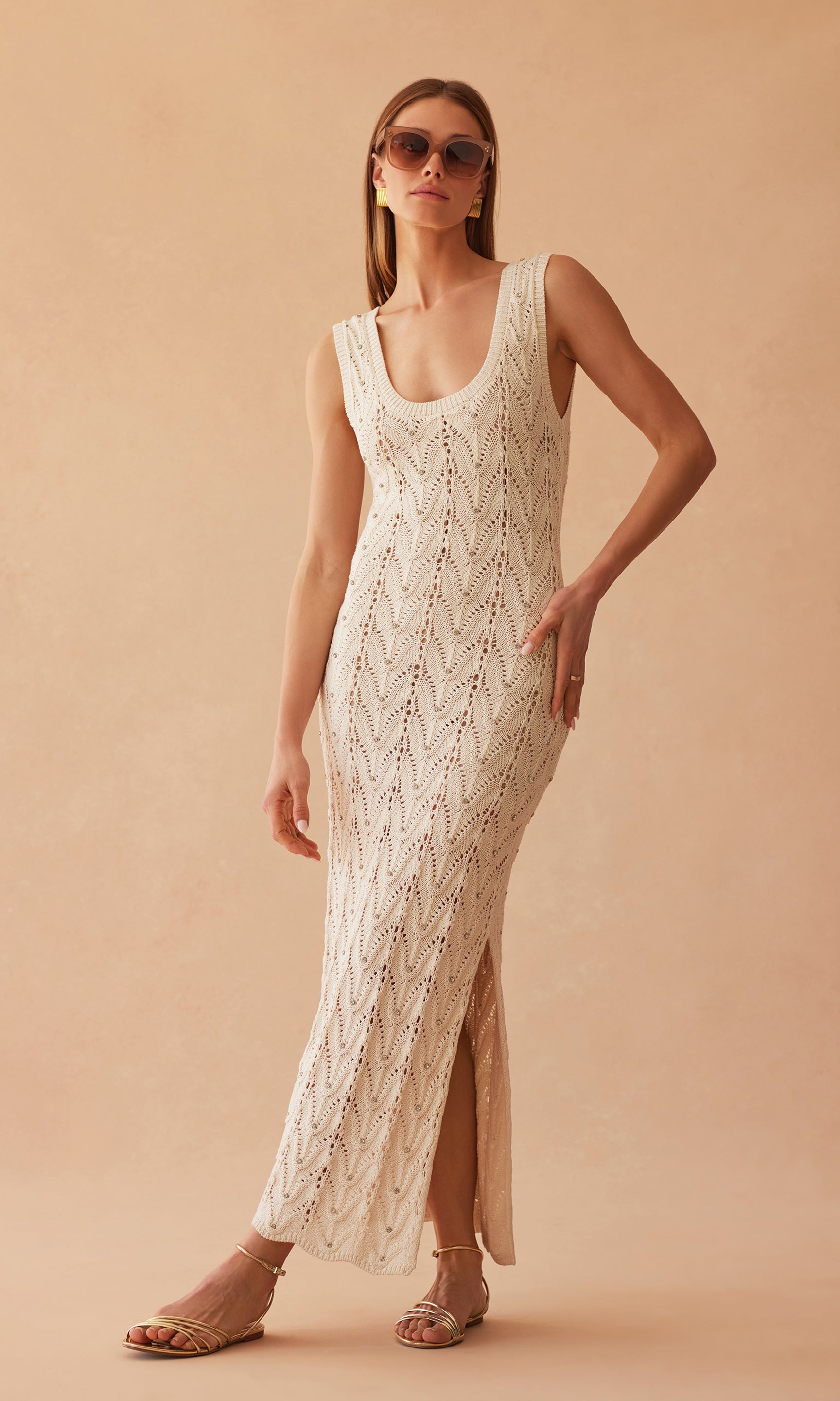 image of model standing in front of a sandy peach wall wearing the Vincent crotchet maxi dress with side slits near the hem, styled with sunglasses and silver sandals