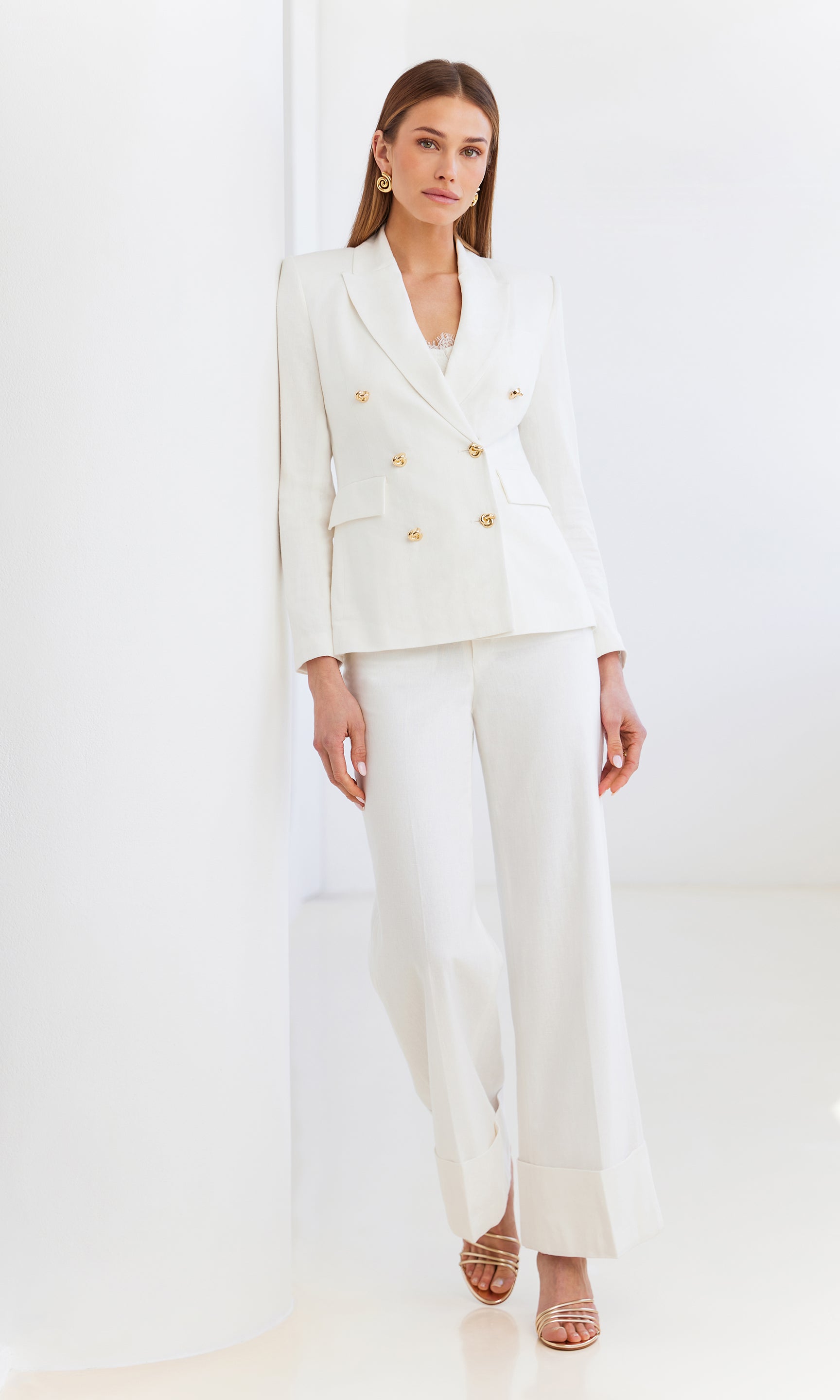 image of model leaning on a white wall wearing the white Arielle blazer with matching white trousers, a white tank and gold earrings